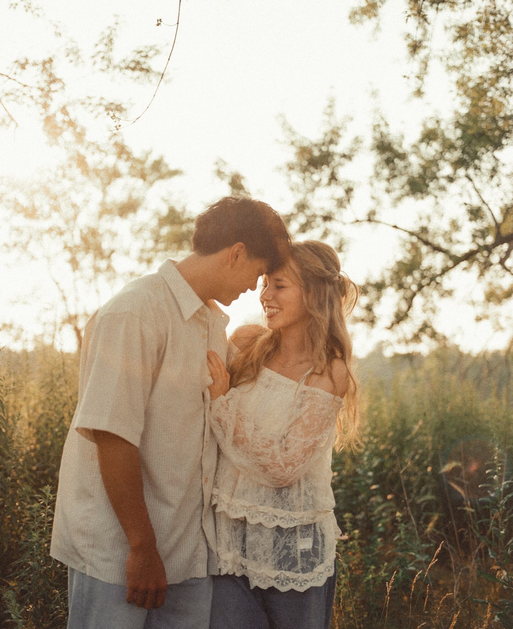 A couple standing close outdoors, smiling, with trees and sunlight in the background.