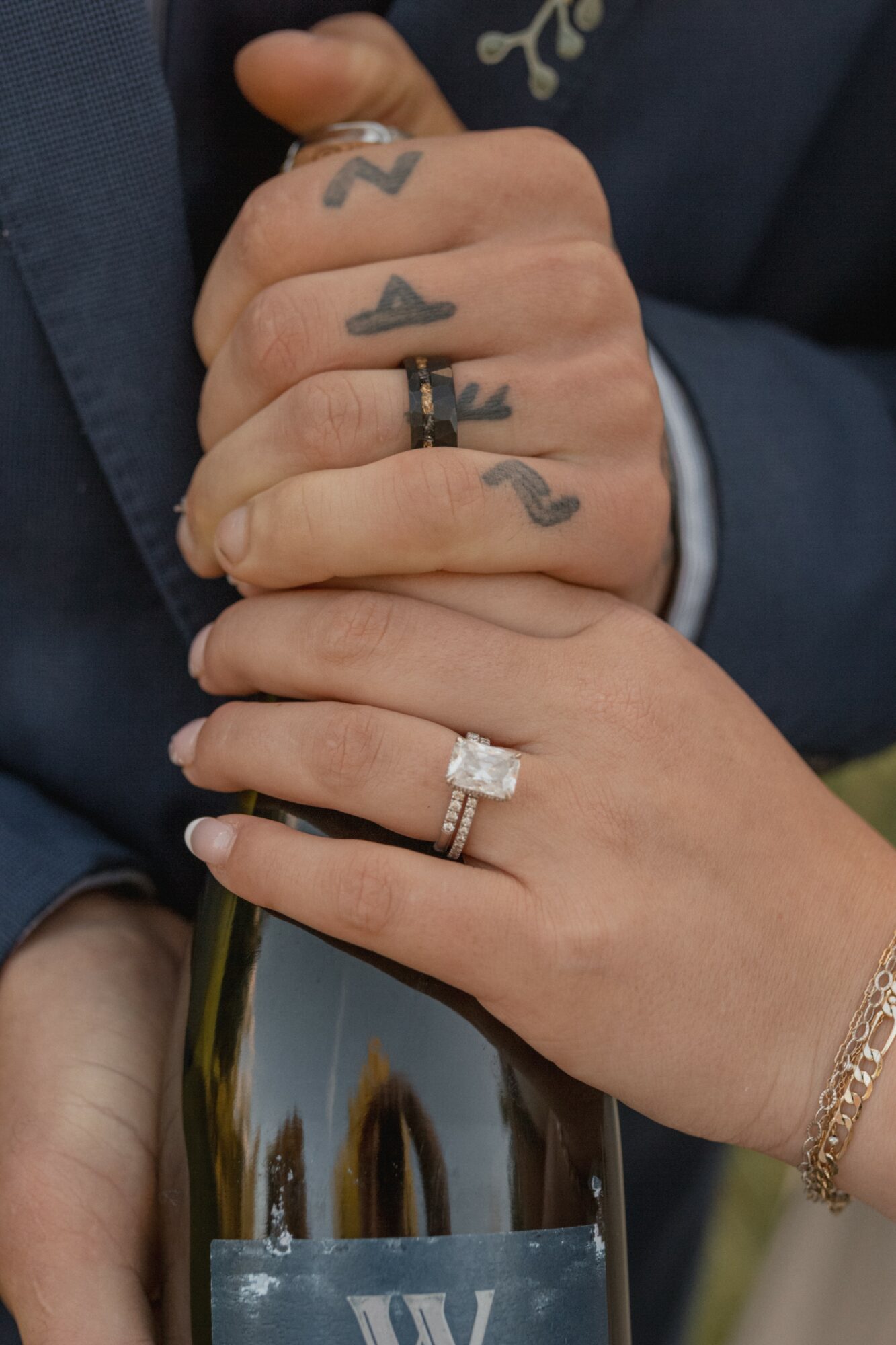 Close-up of a couple's hands with wedding rings, tattoos, and jewelry, holding a bottle, with a blurred outdoor background.