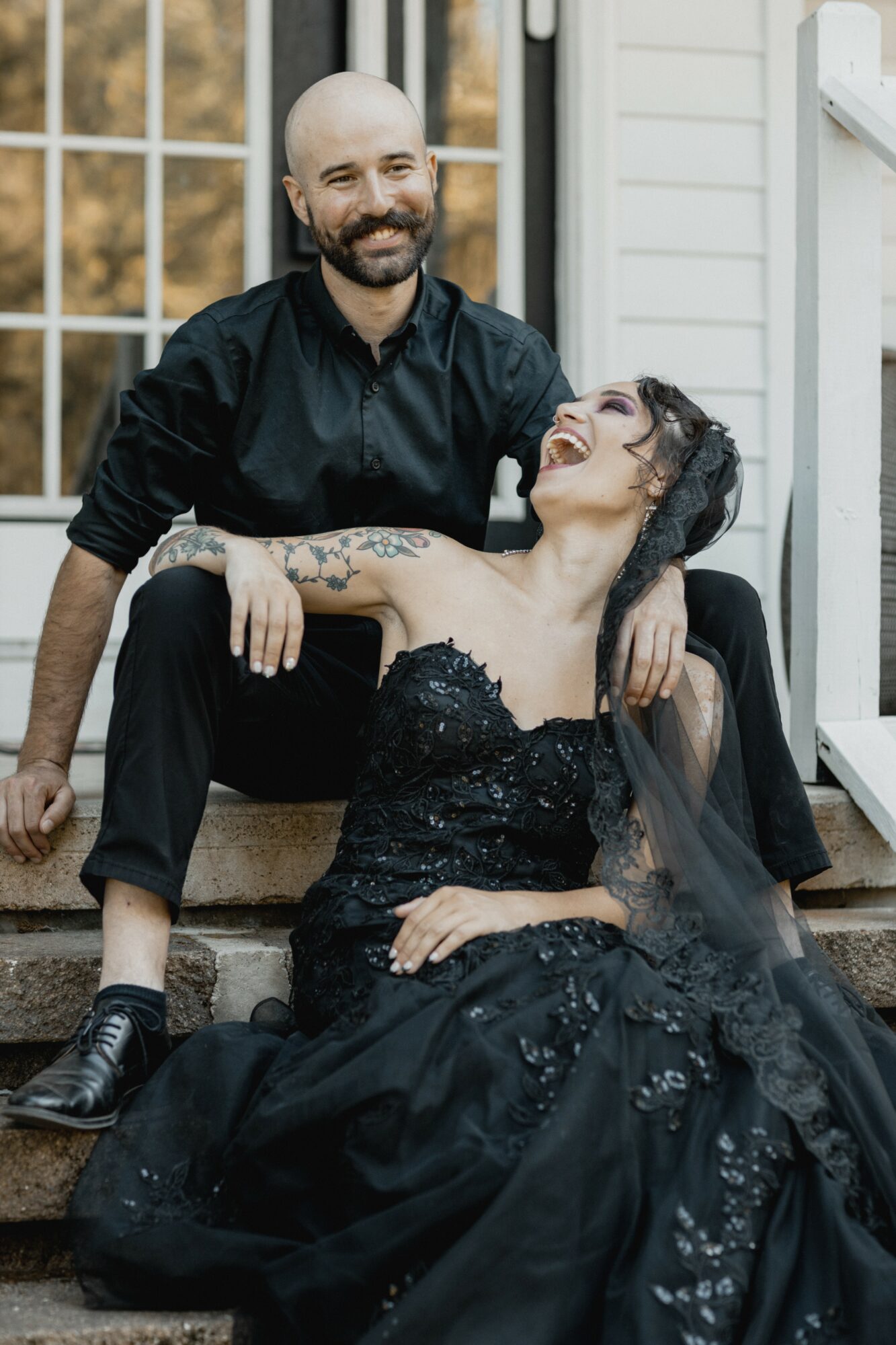 A smiling couple sitting on stairs outside, woman in a black dress and veil, man in black shirt and pants, holding each other.