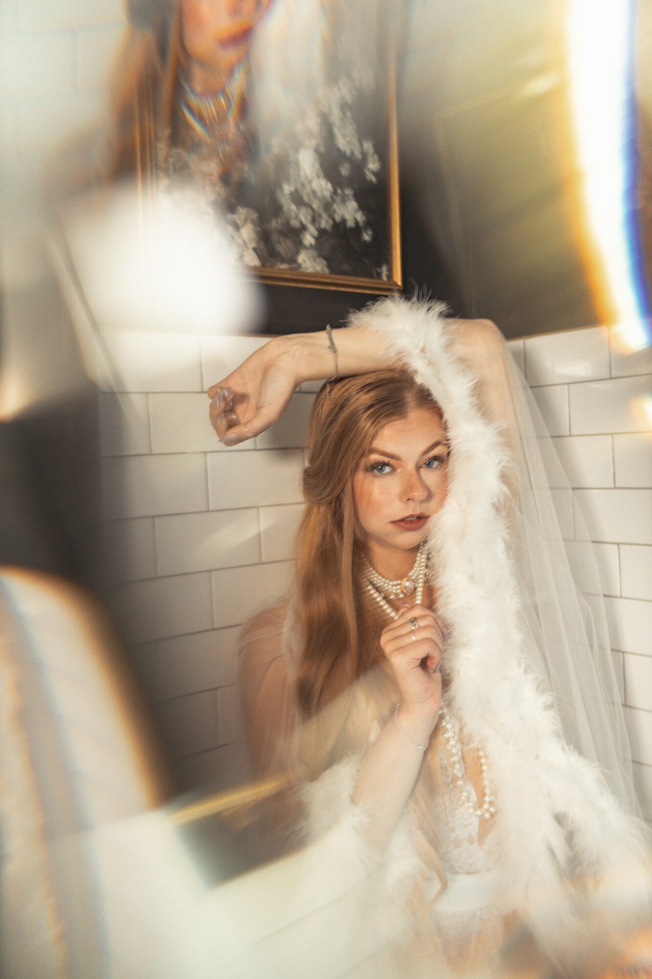 Young woman with long hair and jewelry, posing in front of a mirror with white tiled wall, wearing a white fluffy garment.