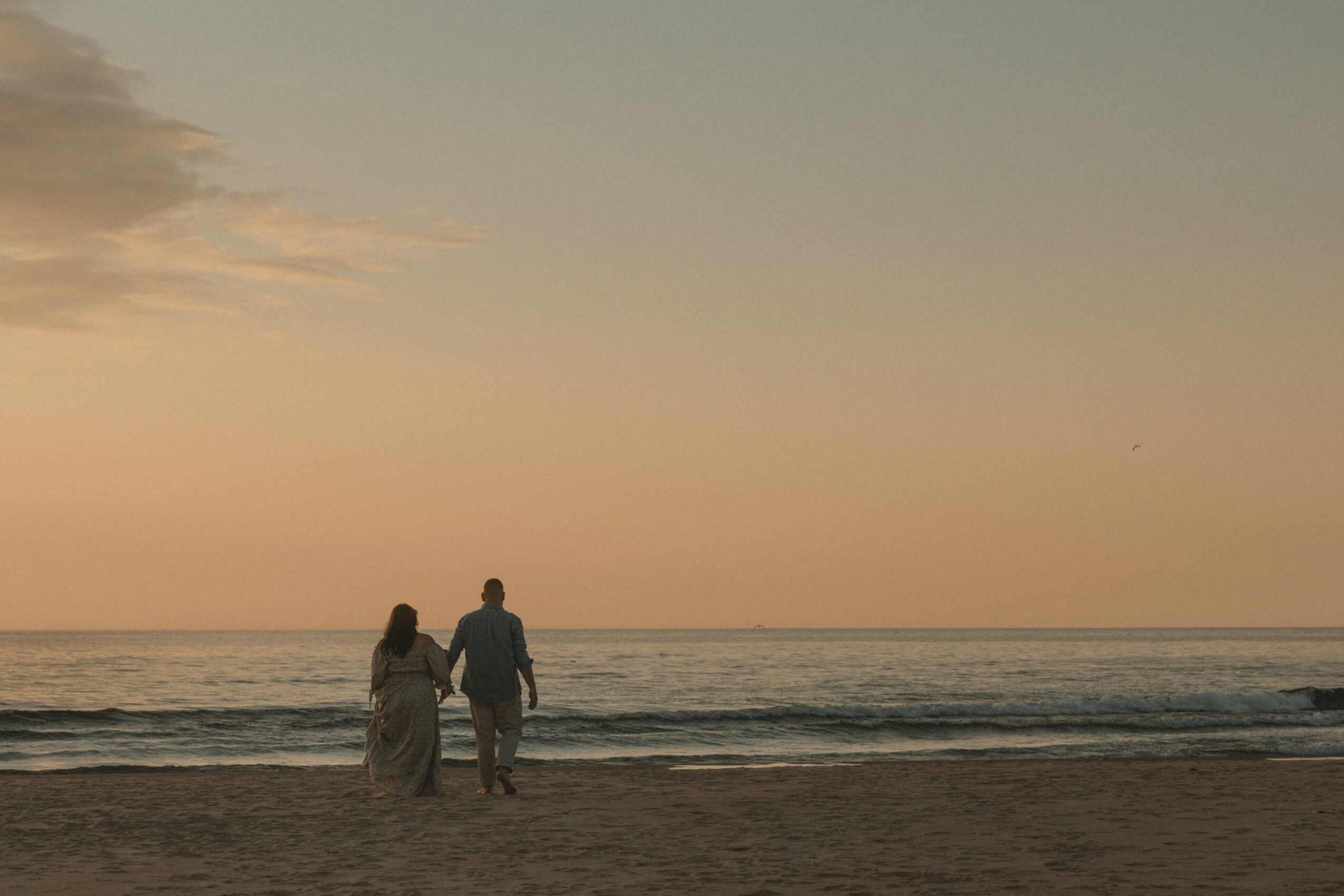 Two people walk along the beach at sunset, holding hands, with the ocean and sky in the background.