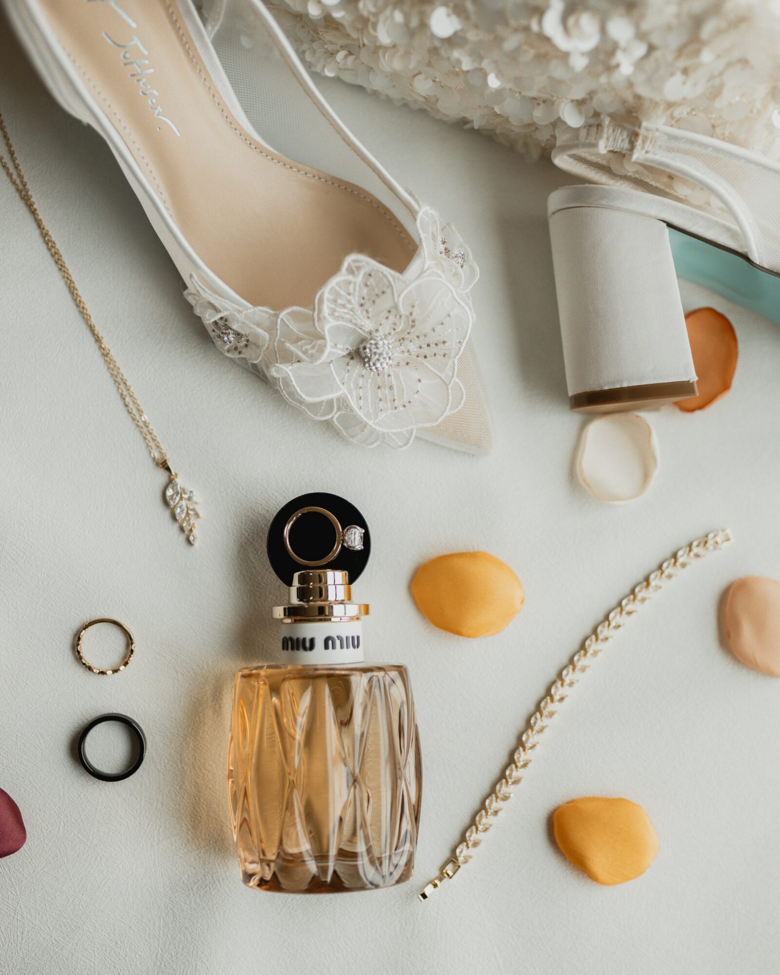 Flat lay of a lace high heel shoe, perfume bottle, jewelry, and scattered flower petals on a light background.