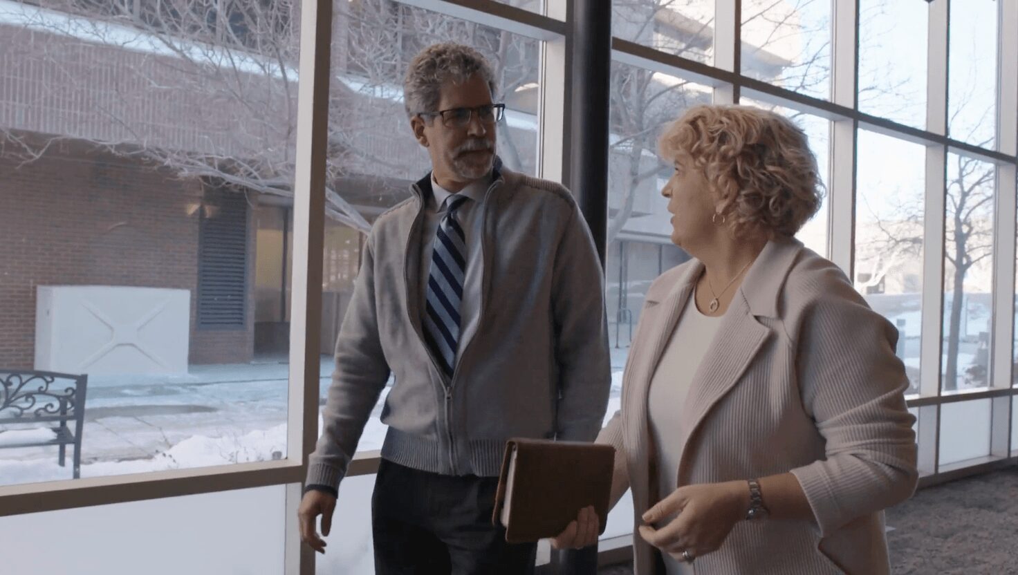 Two people, a man and a woman, are talking indoors near large windows with a snowy scene outside.