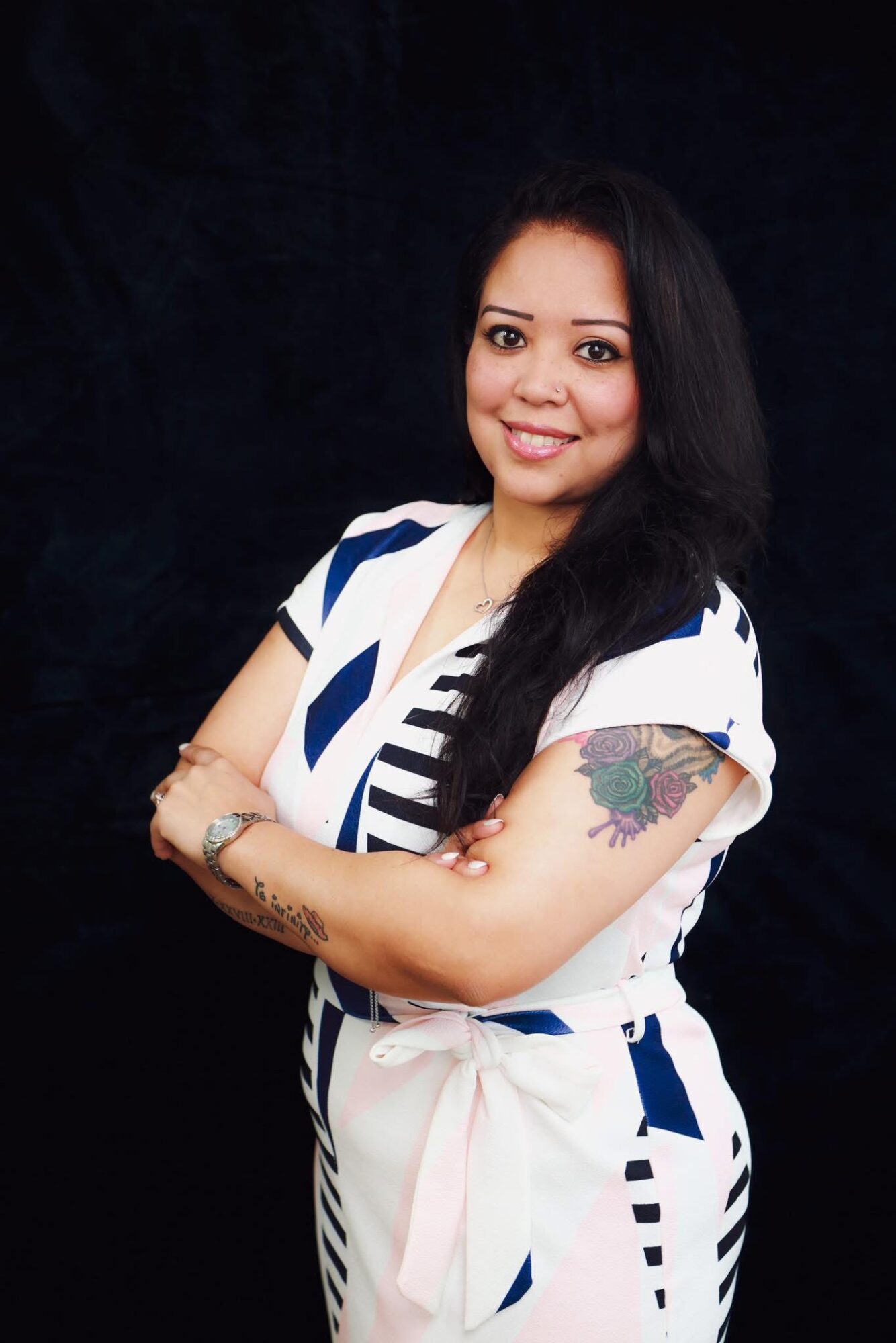 Woman with long dark hair smiling, arms crossed, wearing a white and blue patterned dress, tattoo on her left arm, black background.