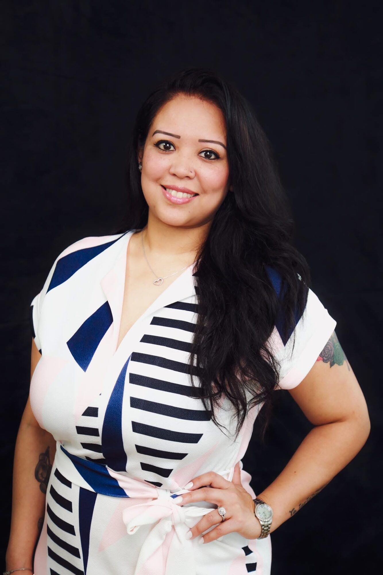 Woman with long dark hair smiling, wearing a white and navy striped dress, standing against a dark background.
