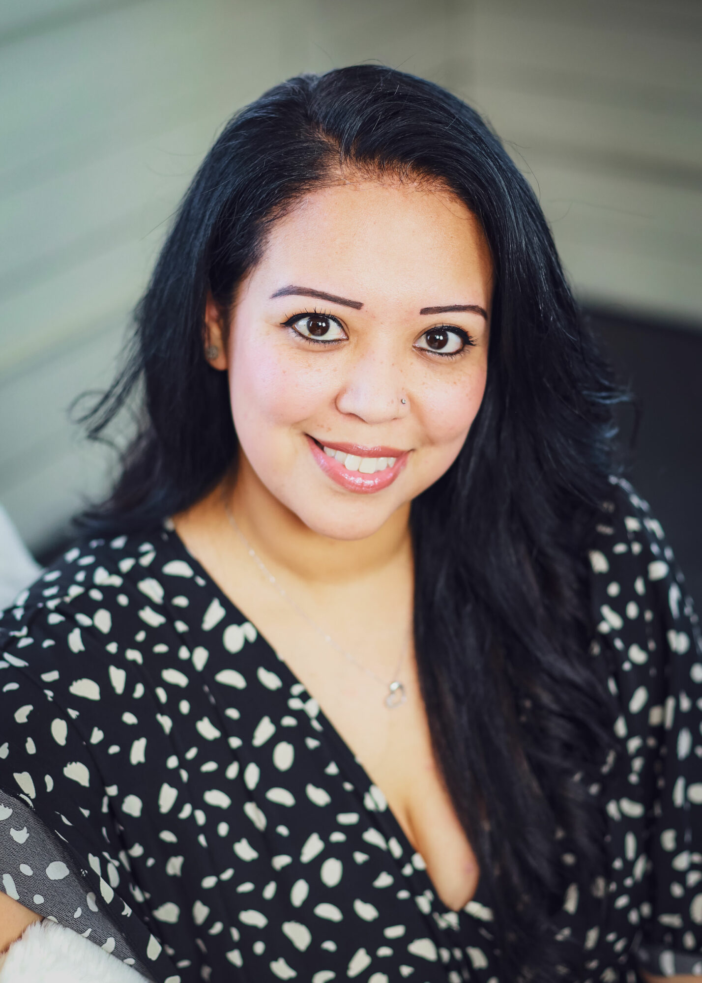 Smiling woman with long dark hair wearing a black and white patterned top, indoors.