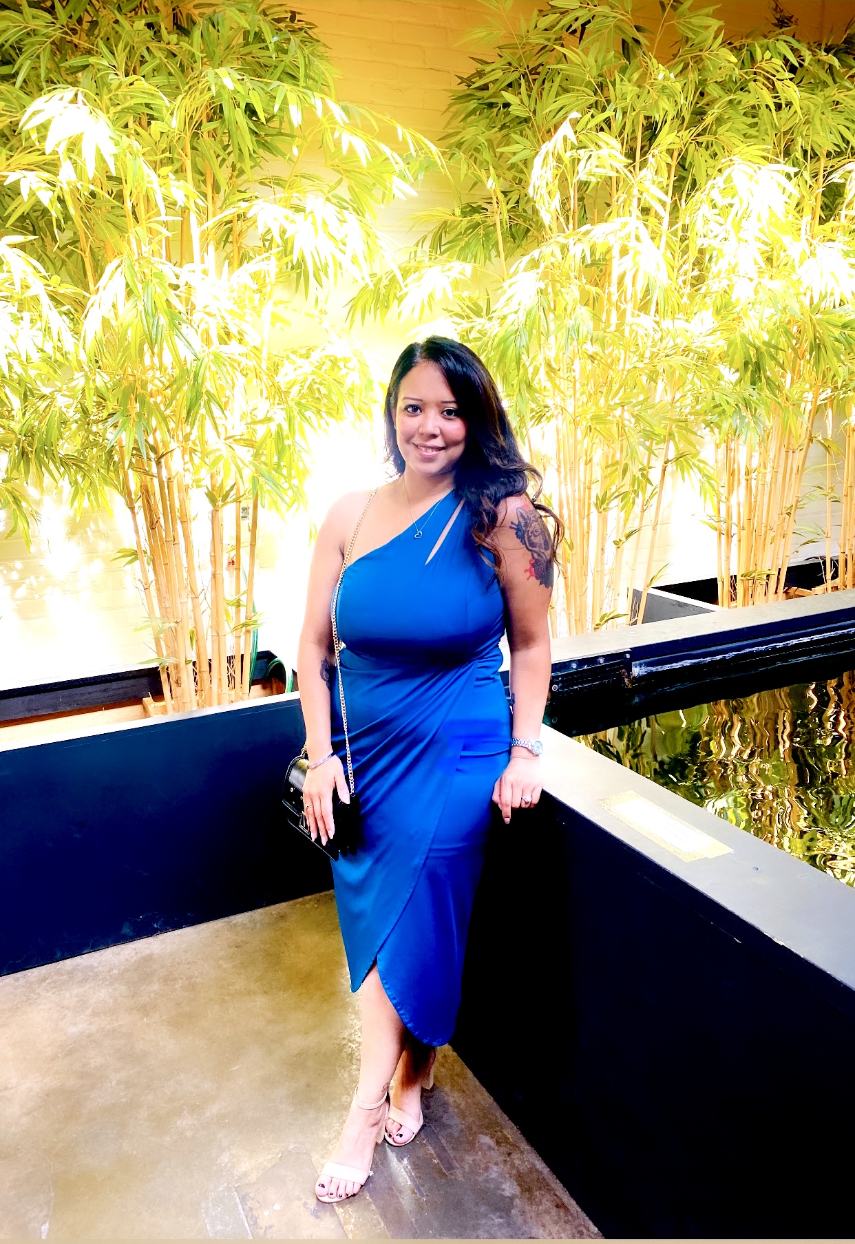 Woman in a blue dress standing on a balcony with green plants behind her, smiling, holding a clutch, wearing heels.