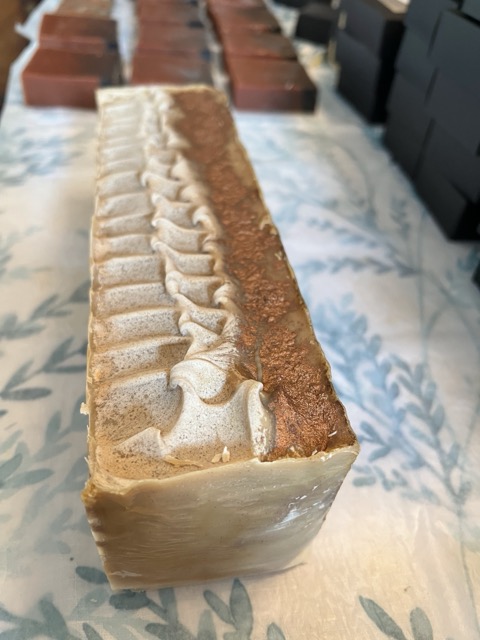 Long rectangular soap bar with textured top, light beige color, placed on a patterned surface with boxes in background.