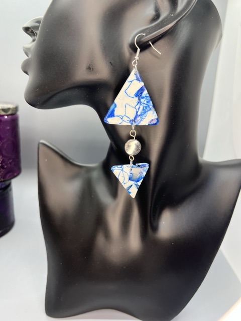 Black mannequin head with a blue and white patterned earring featuring two triangular pieces and a pearl bead, against a light background.