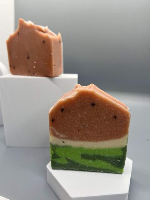 Two layered soap bars with pink, white, and green layers, placed on white blocks against a plain background.