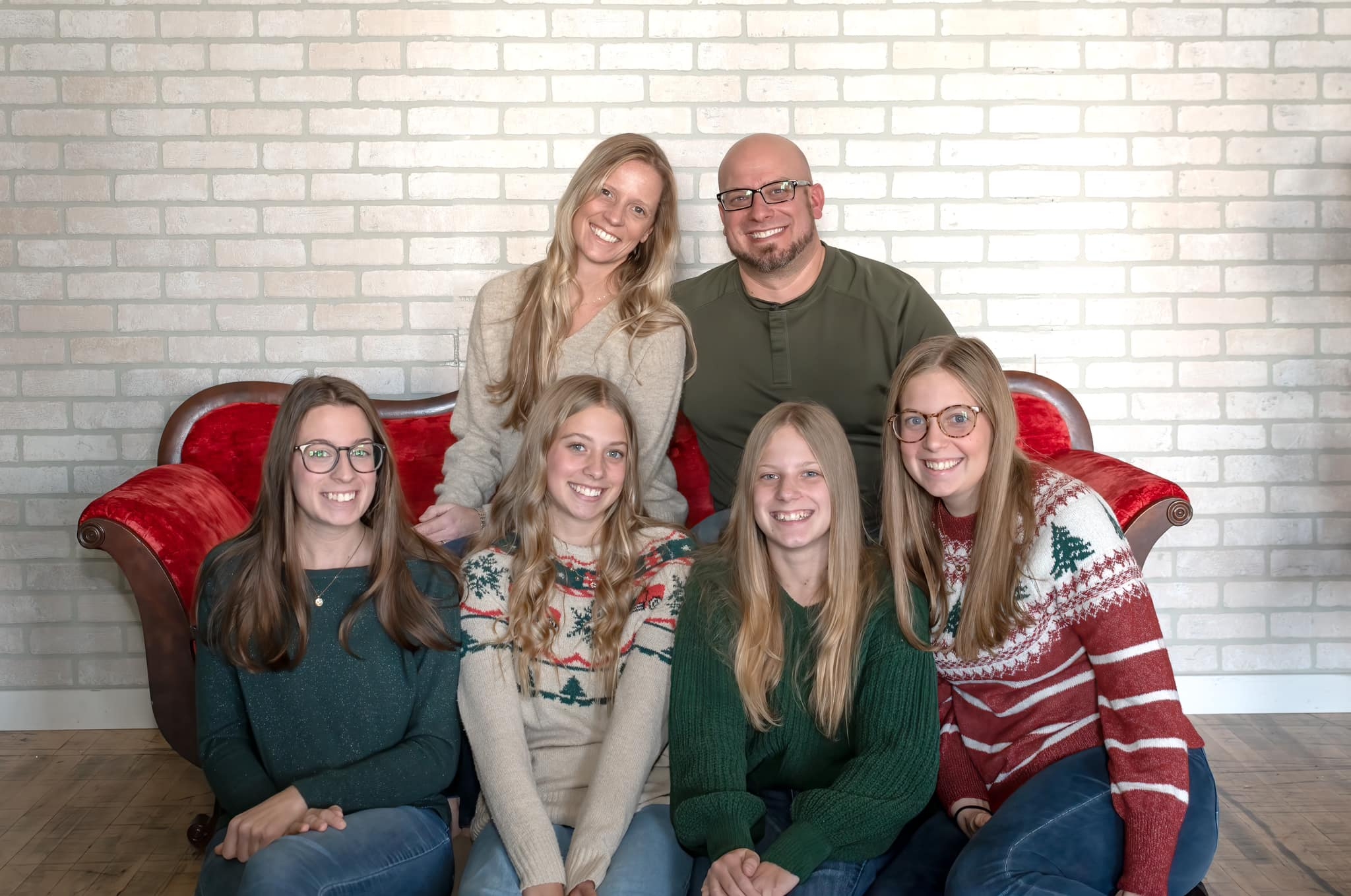 Six people, three adults and three children, sitting on a red couch against a white brick wall, smiling.