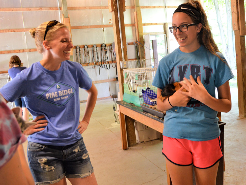 Two young women smiling and talking indoors, one wearing a blue T-shirt and denim shorts, the other in a blue T-shirt and pink shorts.