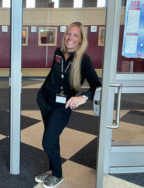 Smiling woman with long blonde hair standing in doorway, wearing black outfit and name badge, in a building lobby.