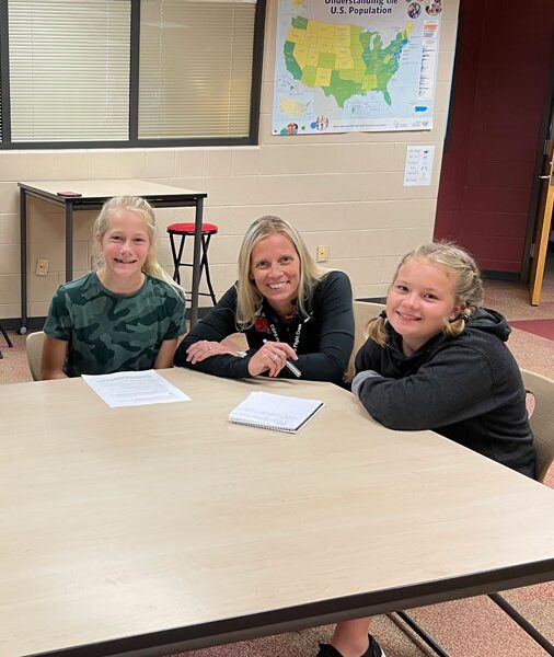Three people sitting at a table in a classroom, smiling, with papers and a map on the wall behind them.