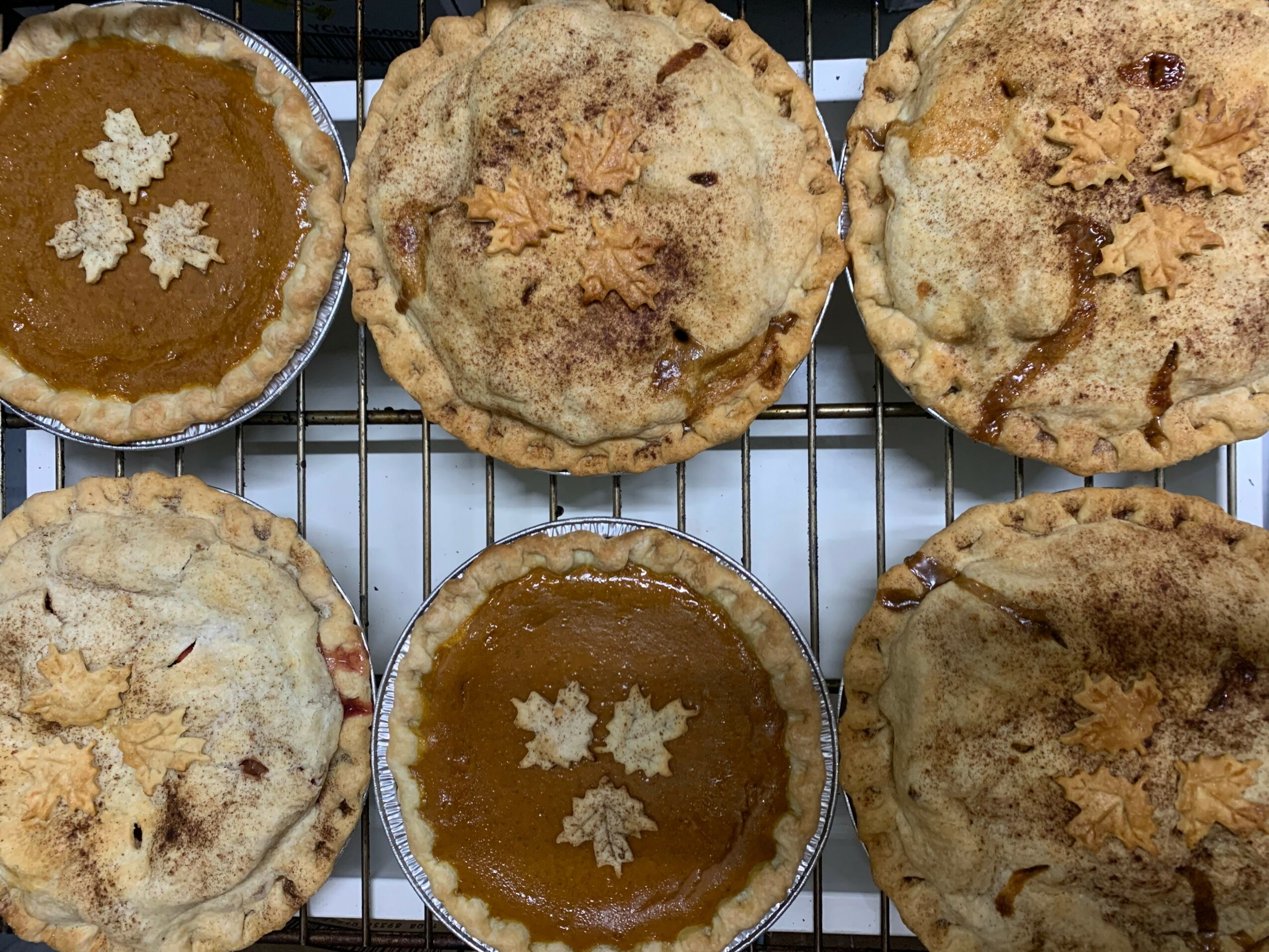 Six pies on a cooling rack, with two filled with caramel and topped with white flowers, and four with crumb toppings.