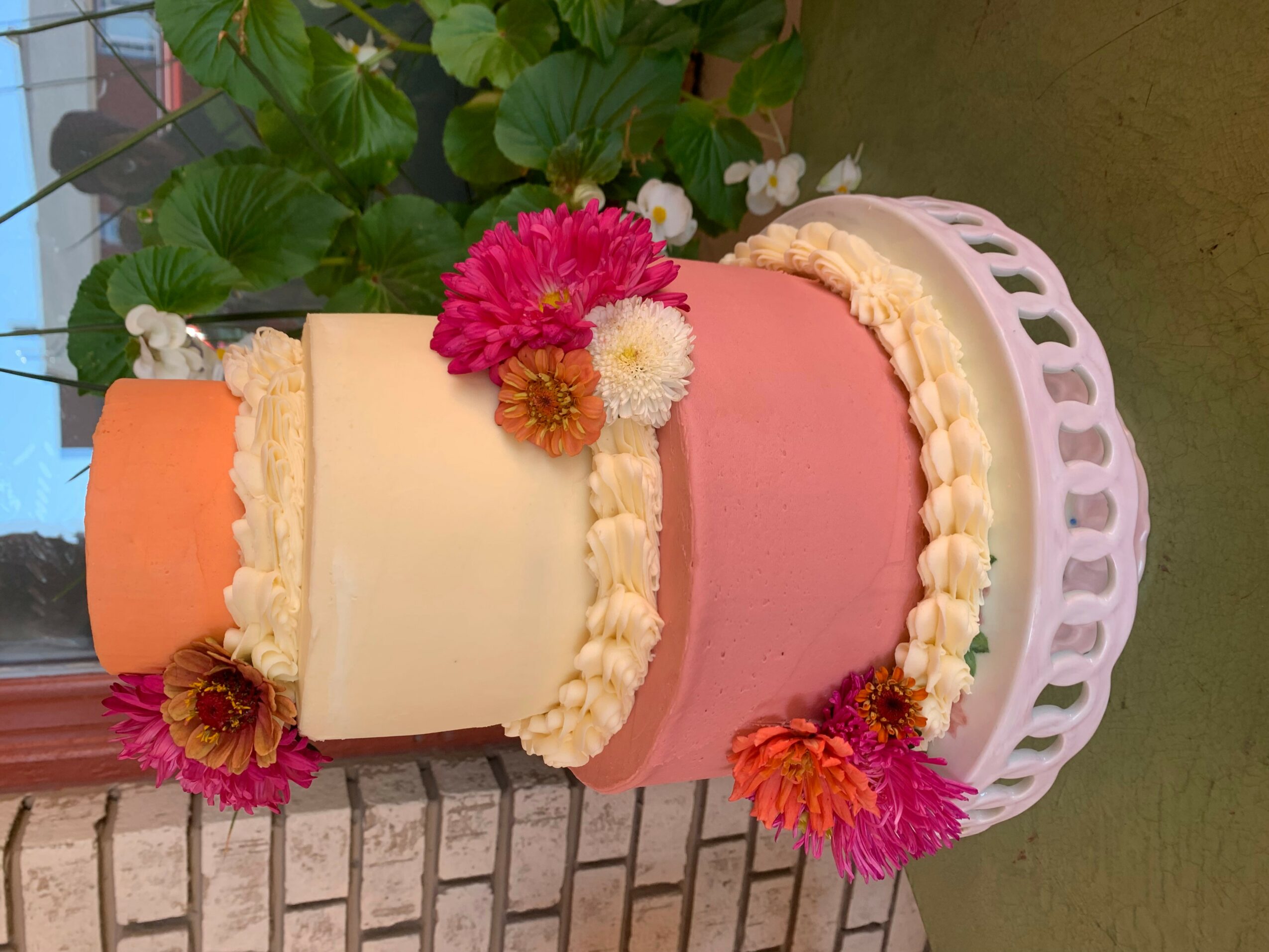 Three-tiered cake with pink, white, and orange layers decorated with flowers, on a white stand, against a brick wall.