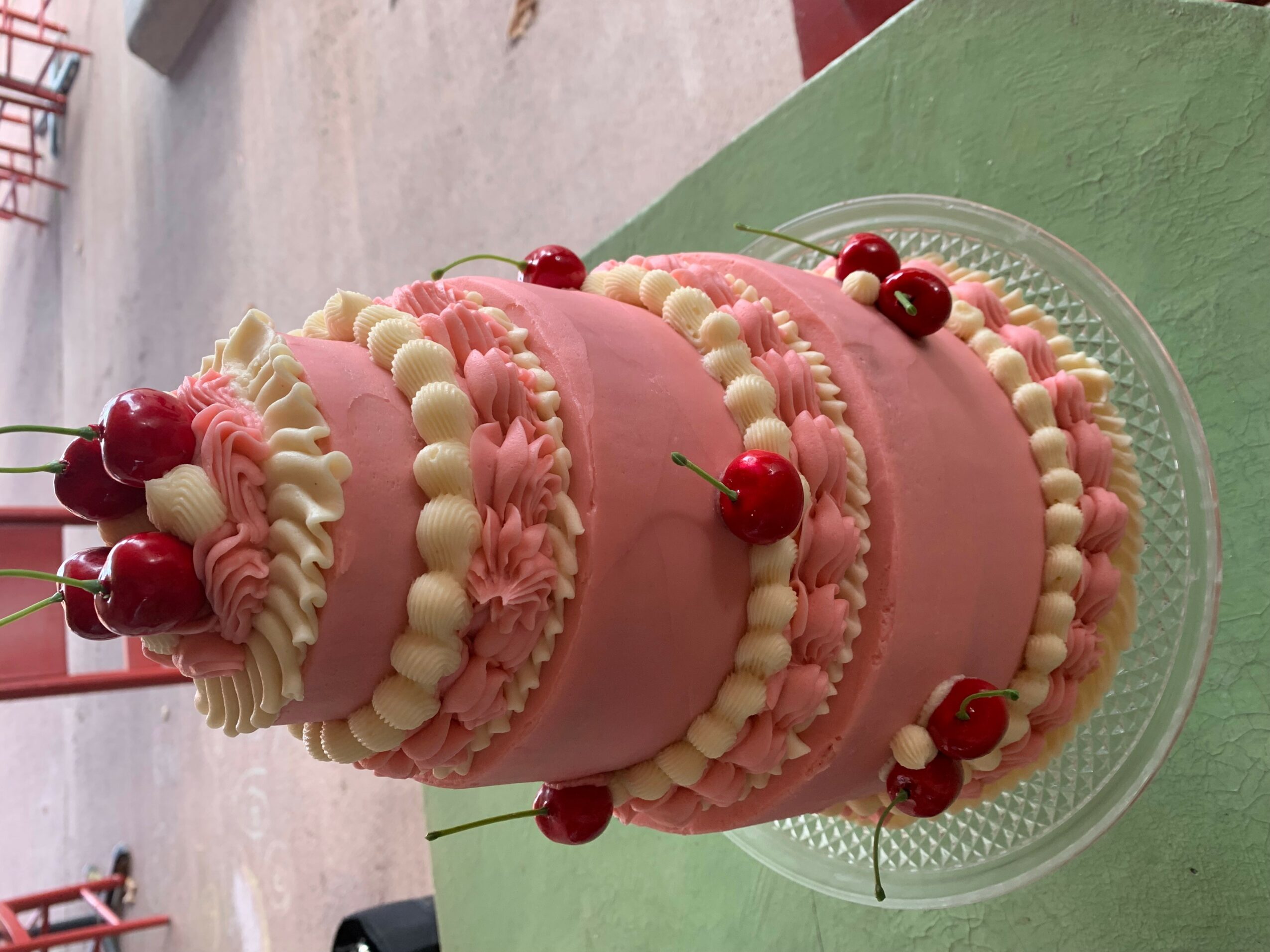 Pink layered cake with white and red cherries, decorated with white cream, on a glass plate.