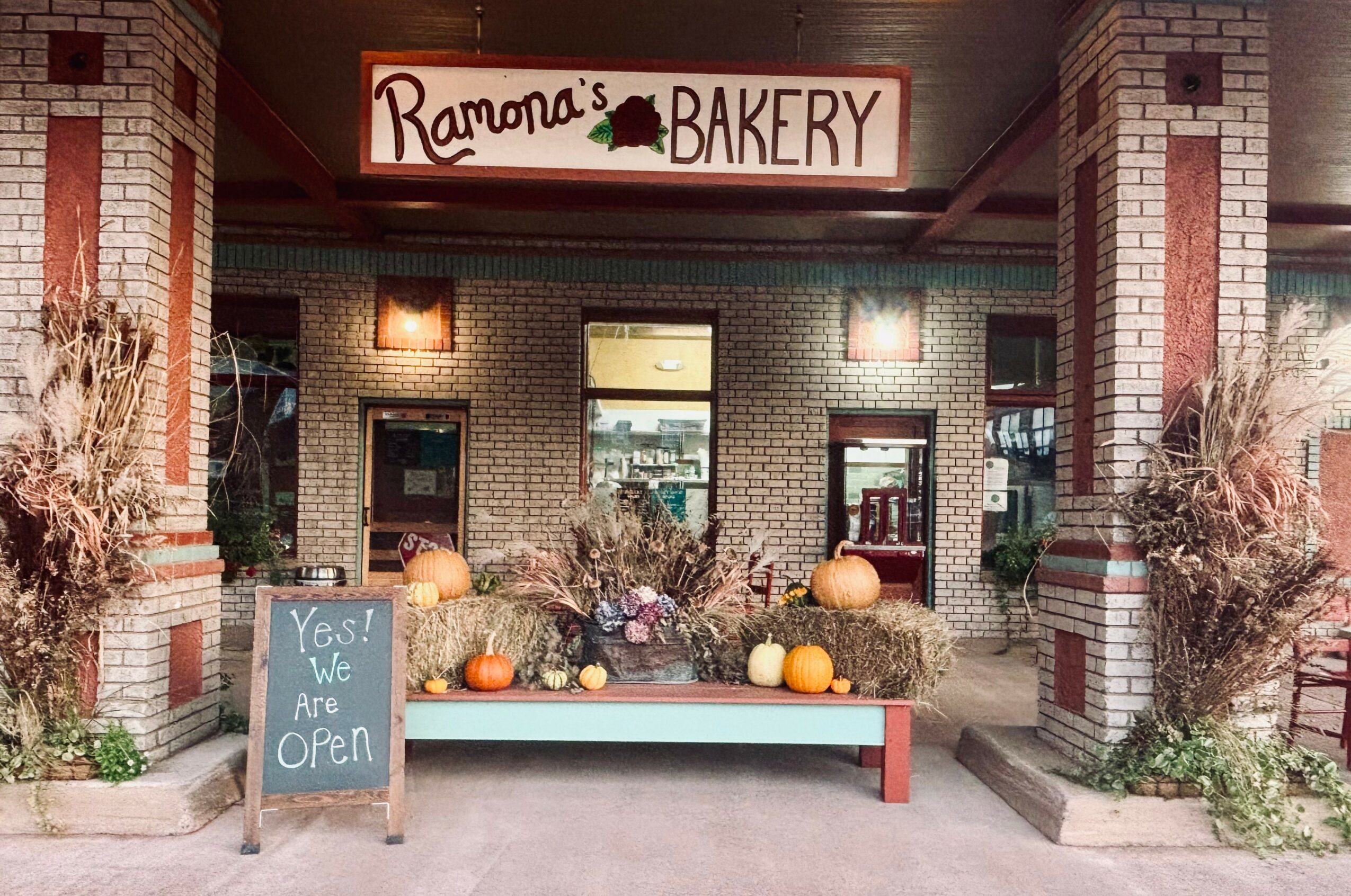 Bakery storefront with pumpkins and dried plants outside, sign reading 'Ramona's Bakery,' and a chalkboard sign saying 'Yes! We Are Open.'