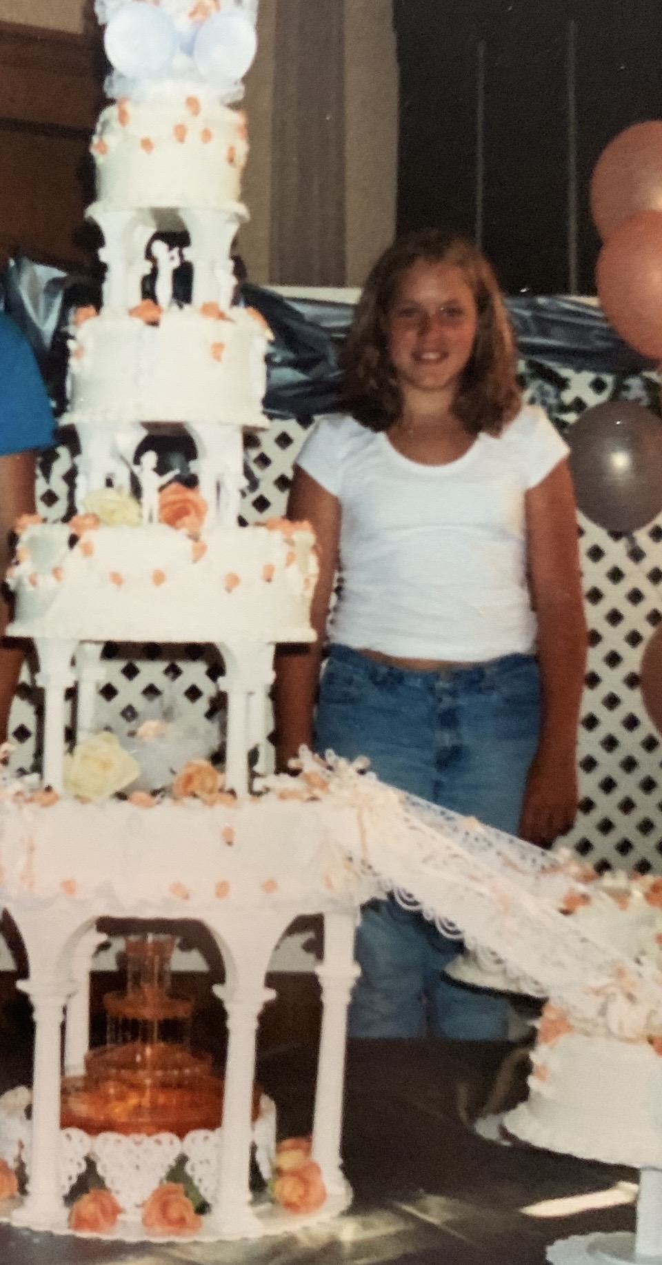 Girl with curly hair in white shirt and jeans standing next to a multi-tiered cake with white lattice and orange decorations.