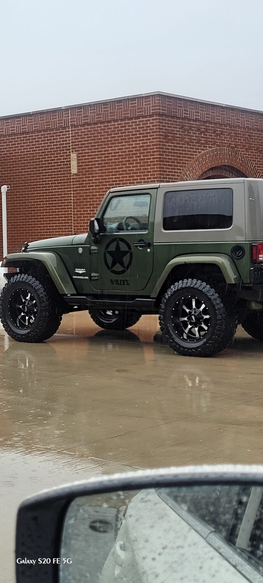 Green vehicle parked on wet ground near a brick building, with part of a silver car visible in the foreground.