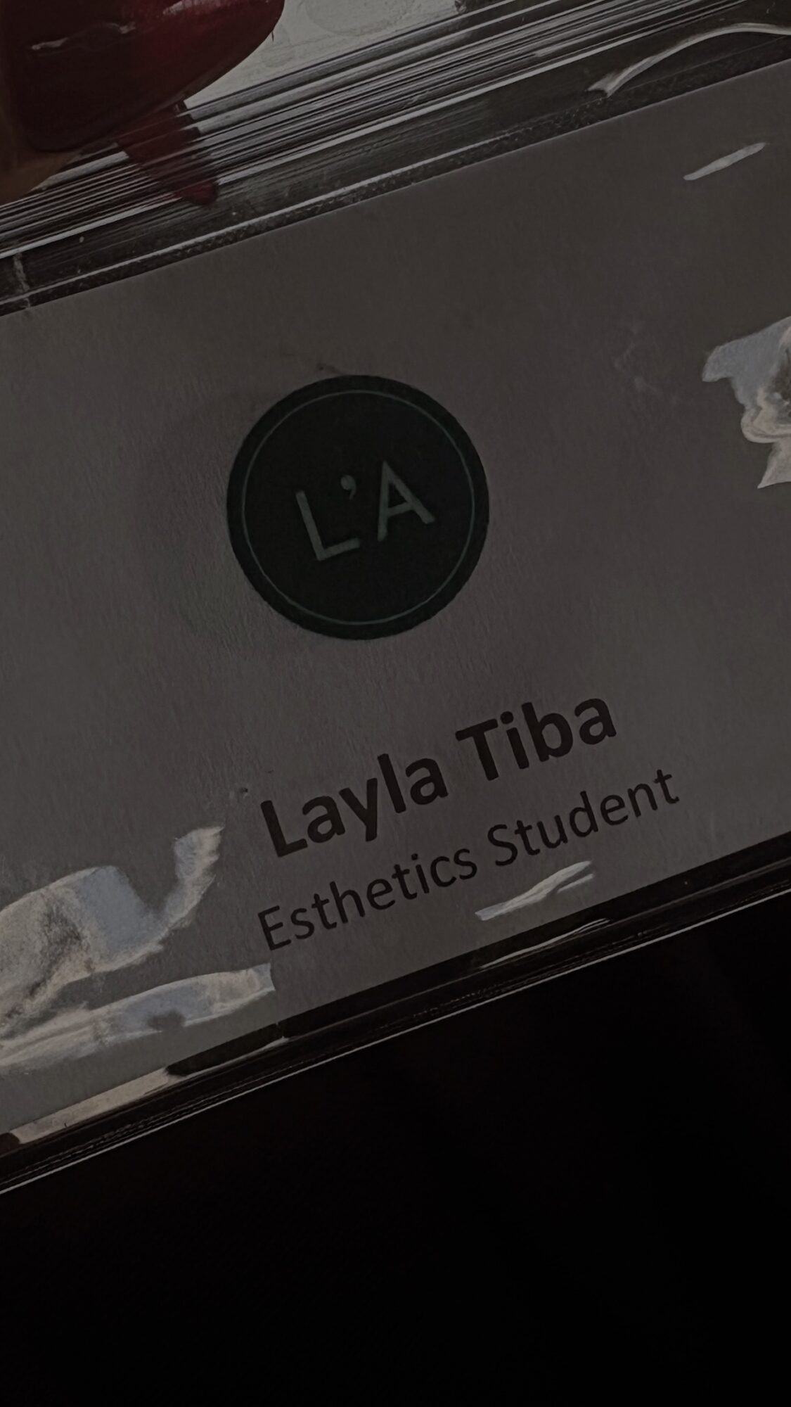 Name tag with initials 'L.A' and name 'Layla Tiba', 'Esthetics Student' title, on clear plastic surface.