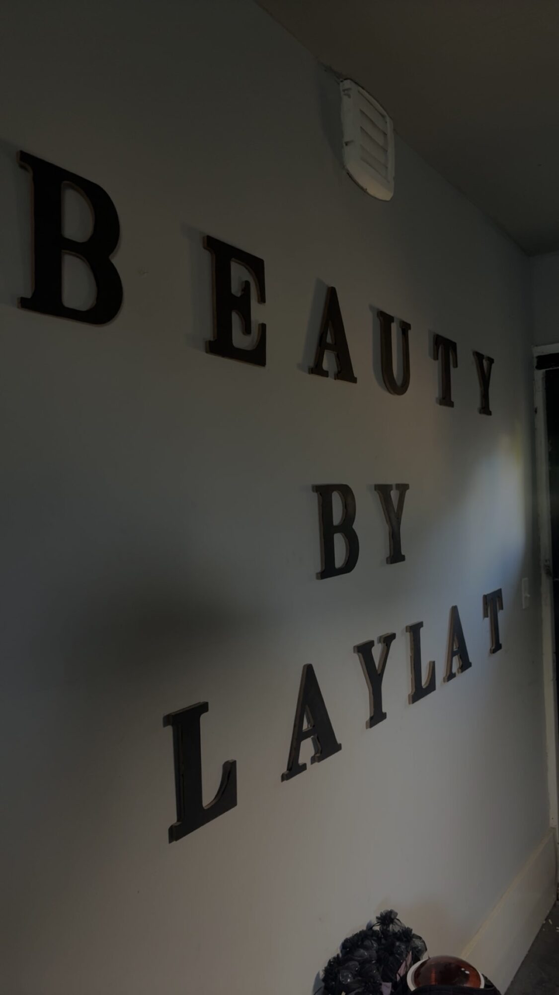 Wall with black letters spelling 'BEAUTY BY LAYLA' arranged vertically, part of a room interior, with a window on the right.