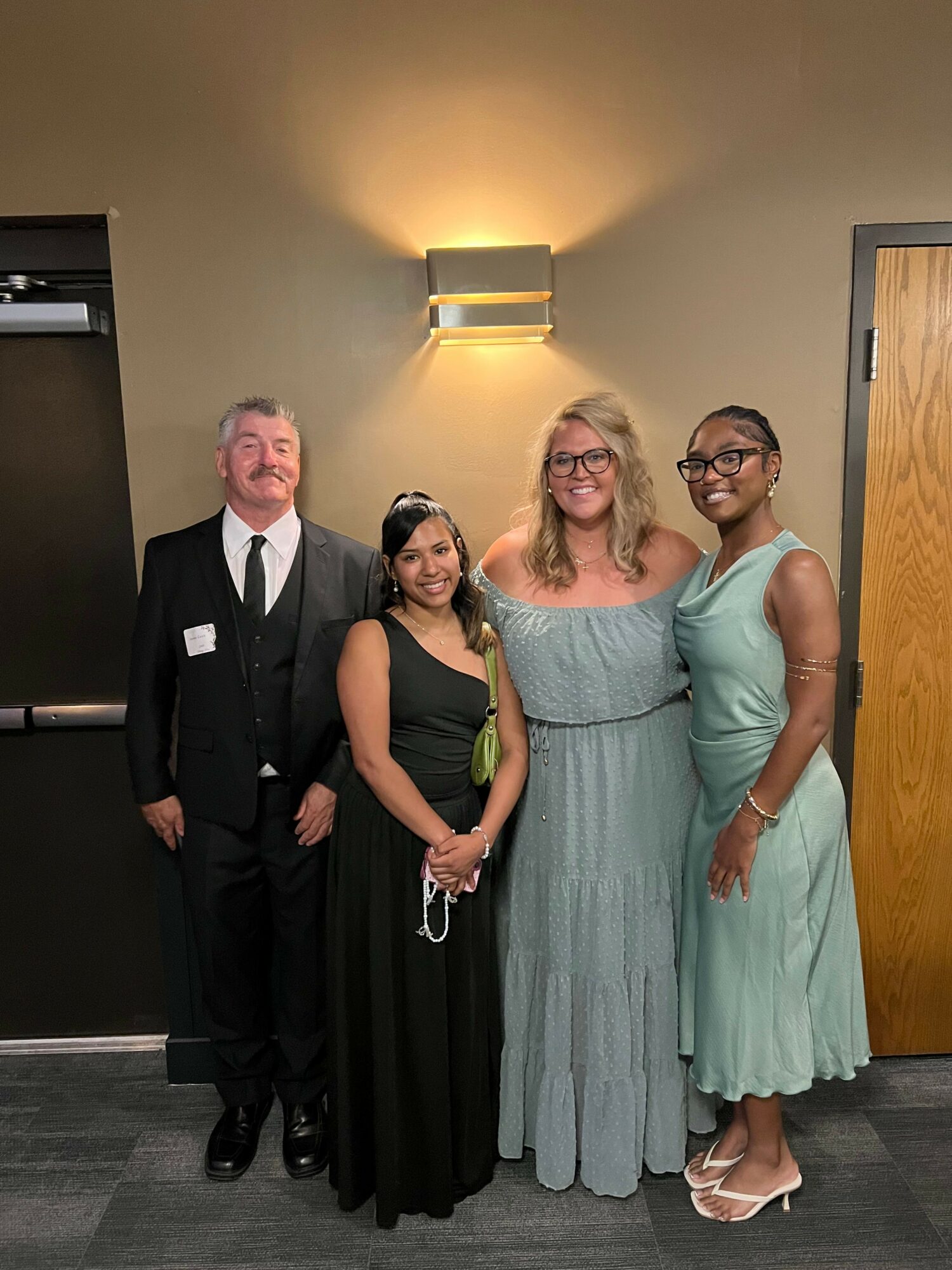 Four people standing together indoors, smiling, with a wall and door in the background.