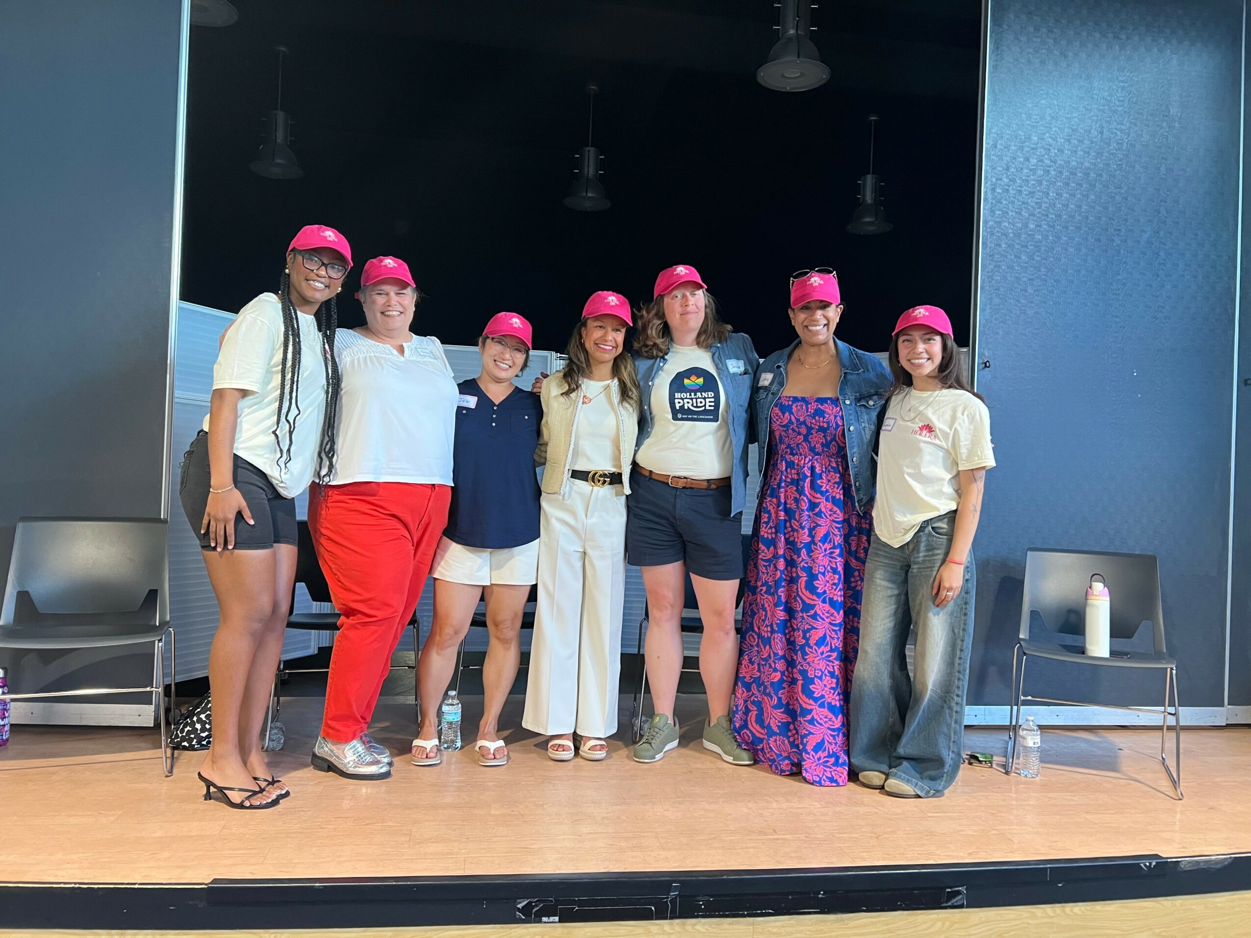 Seven people stand on a stage, wearing pink hats and casual clothing, with chairs and a candle in the background.