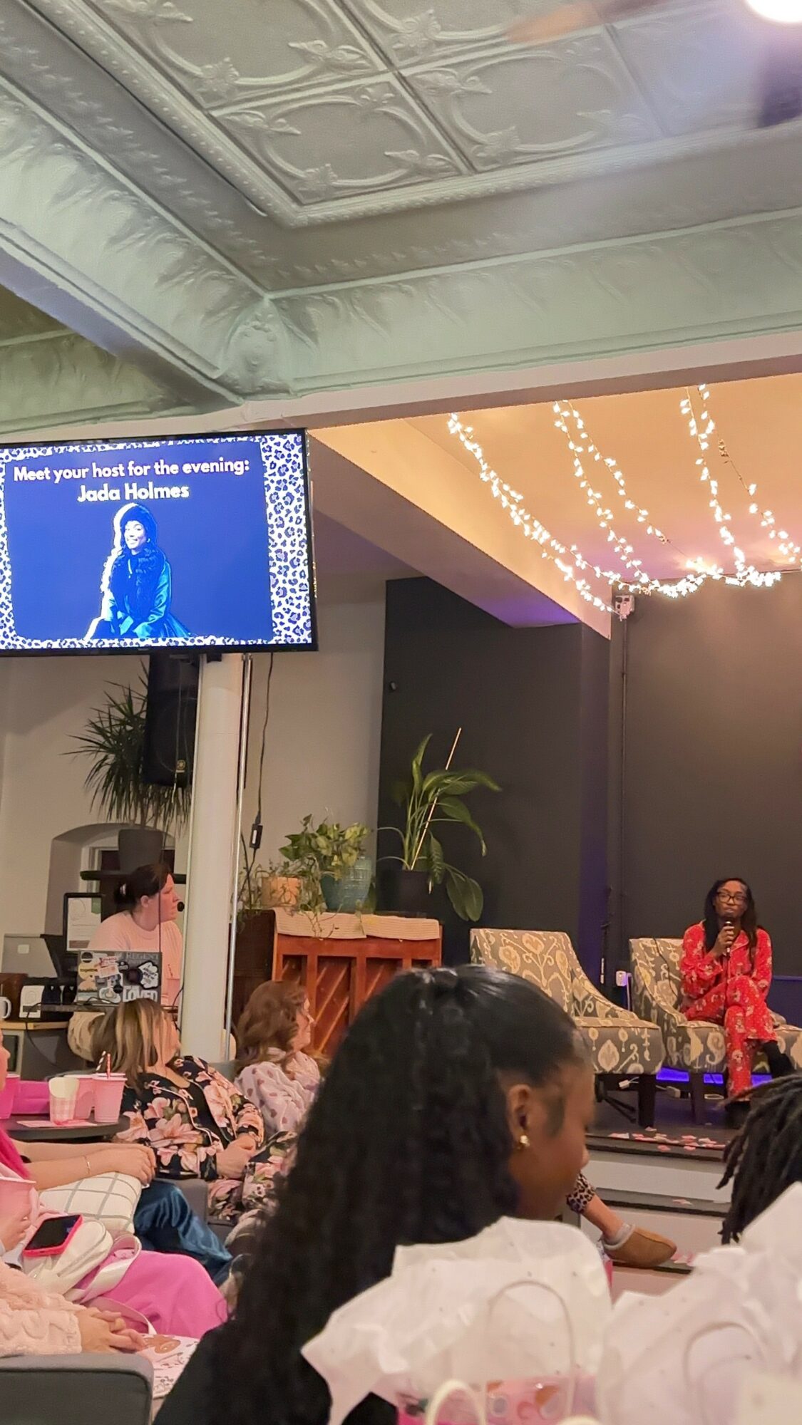 People seated in an indoor event space with a stage, a woman speaking, and decorative lighting on the ceiling.