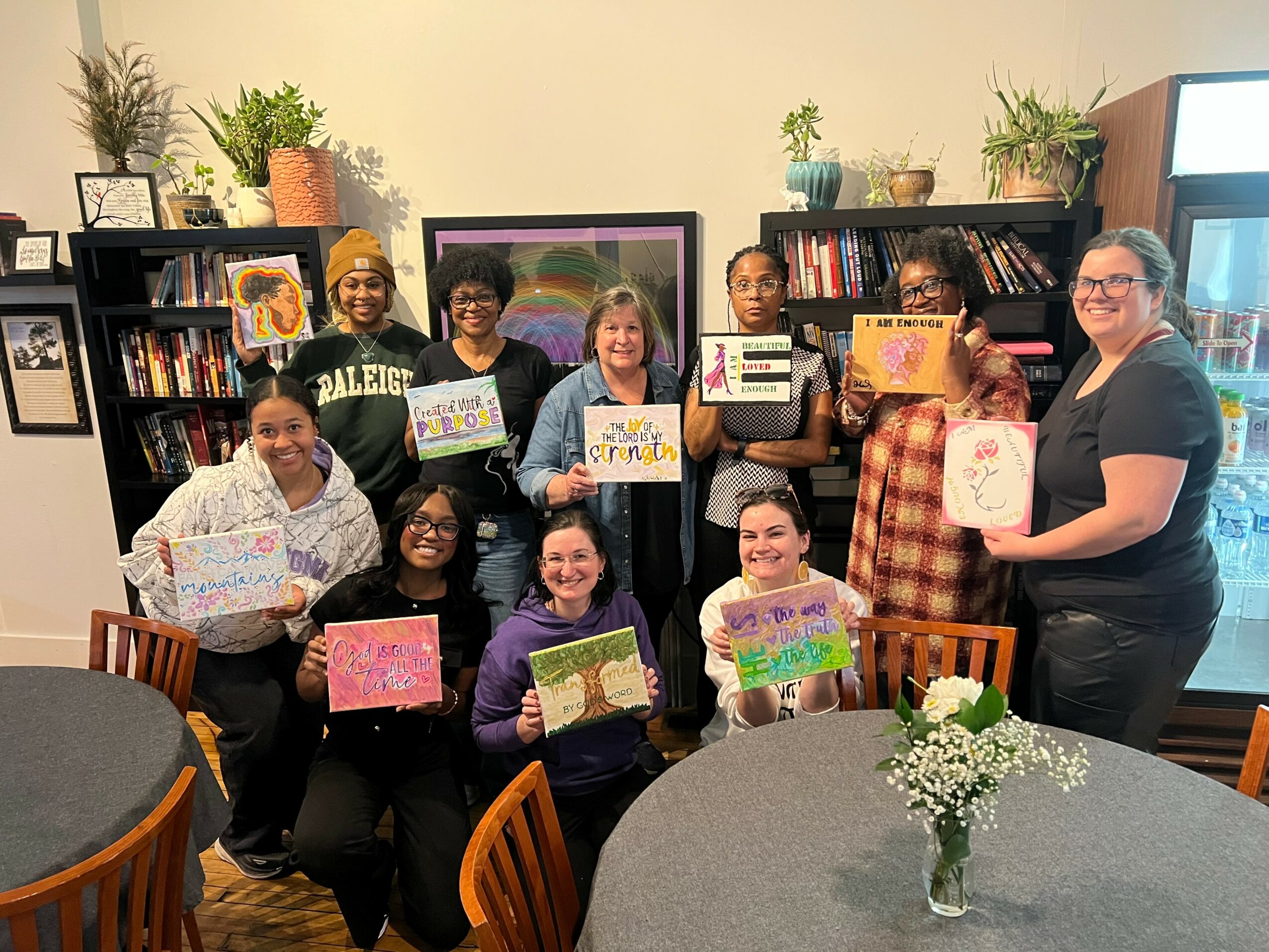 Group of ten people holding colorful signs in a room with bookshelves and framed artwork, smiling at the camera.