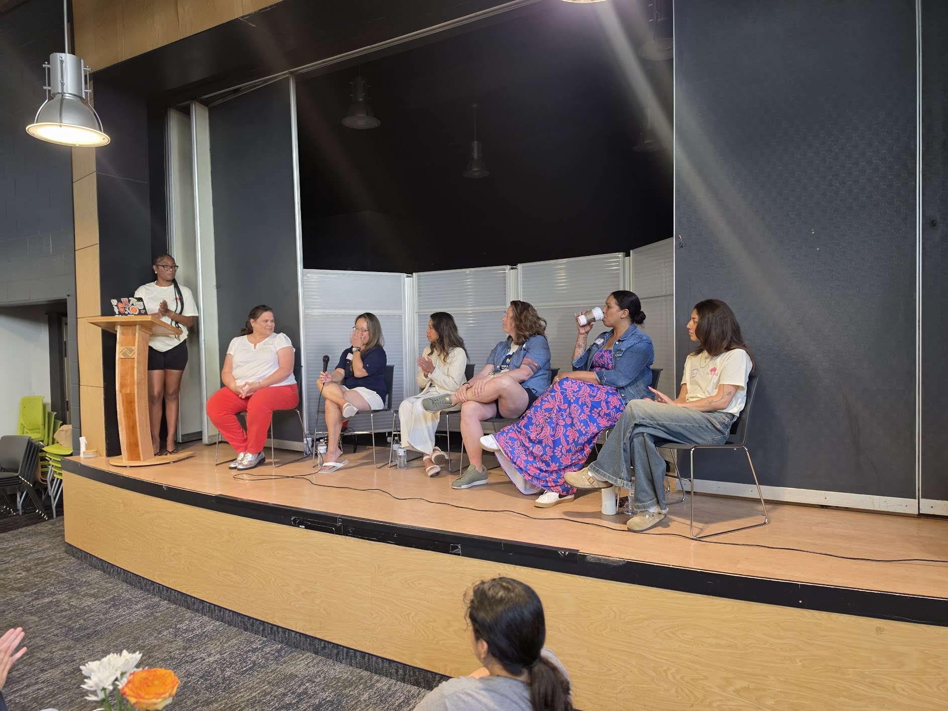 Panel of seven people seated on stage, one person standing at a podium, in a conference room.