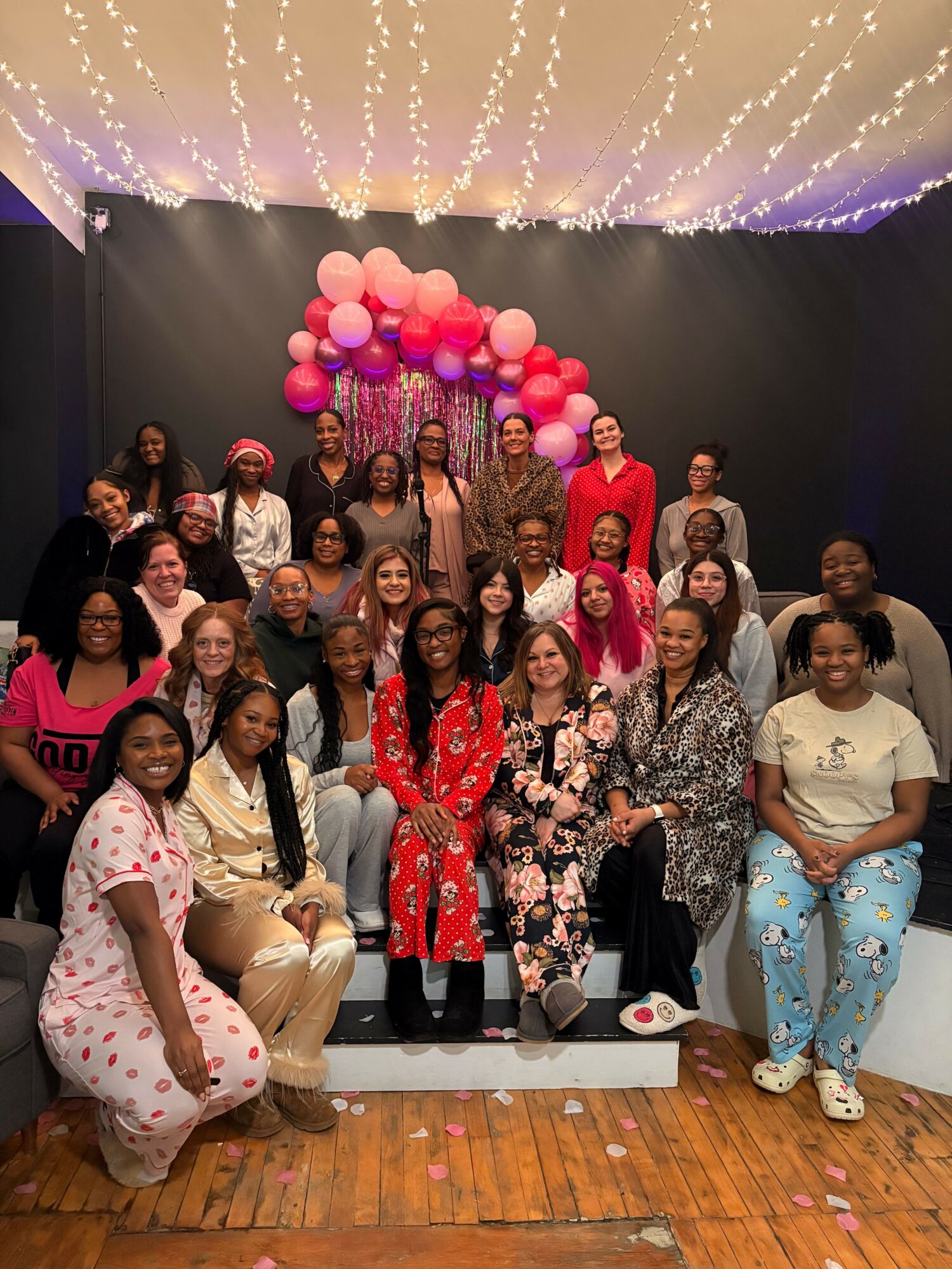 Group of women and girls posing indoors with pink balloons and string lights overhead.
