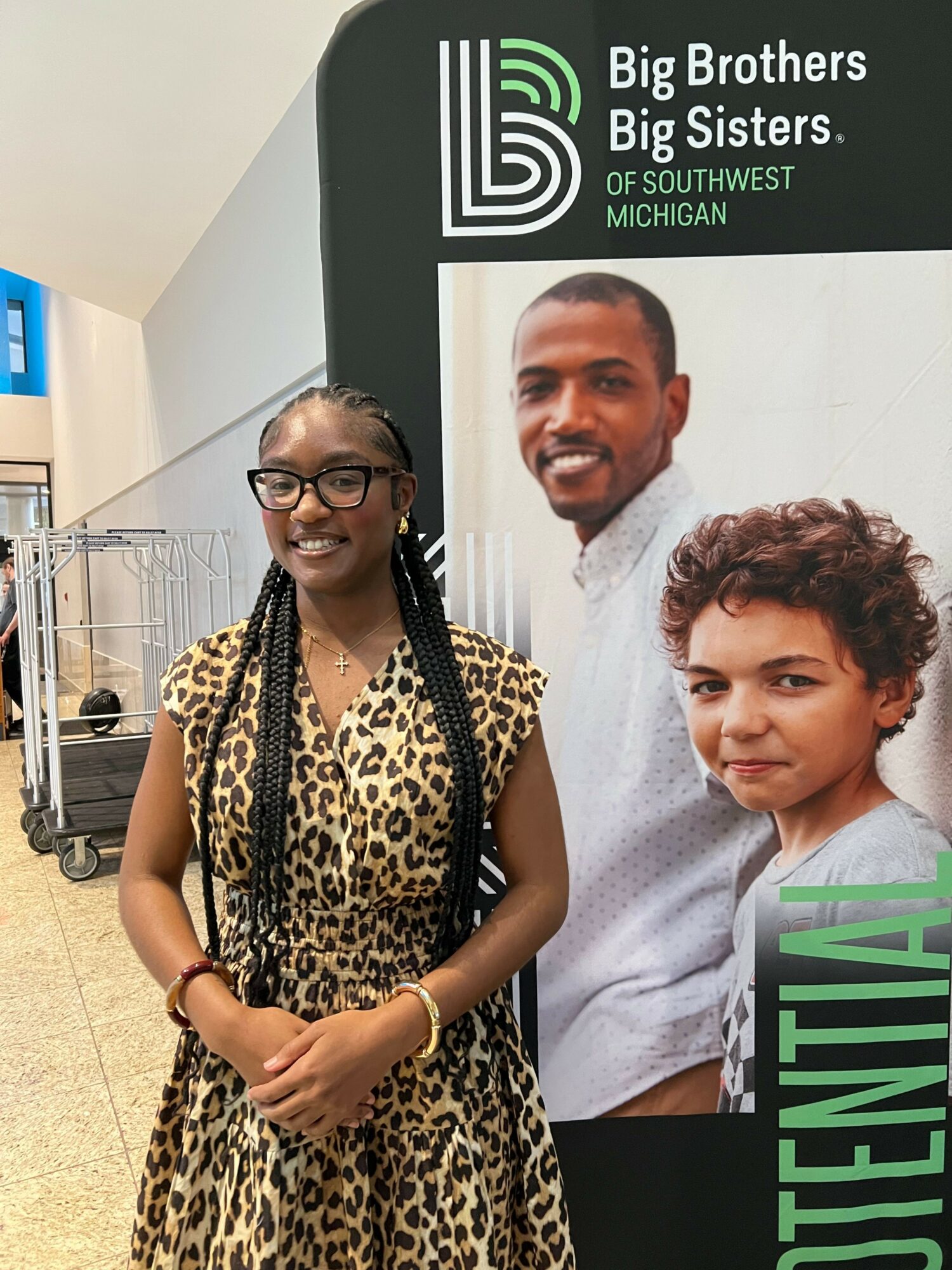 Young girl with glasses and braided hair standing next to a large poster of two men and a boy, smiling.
