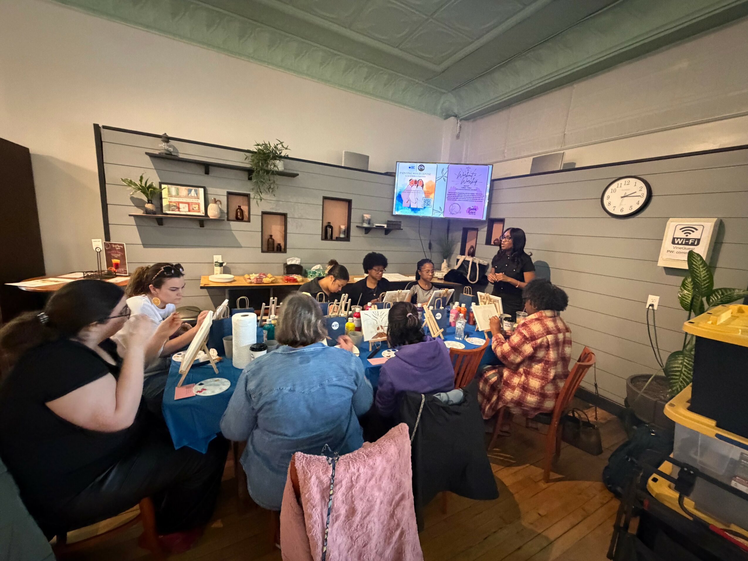 Group of people sitting around a table in a room with shelves, a TV, and a clock on the wall, engaging in a meeting or discussion.