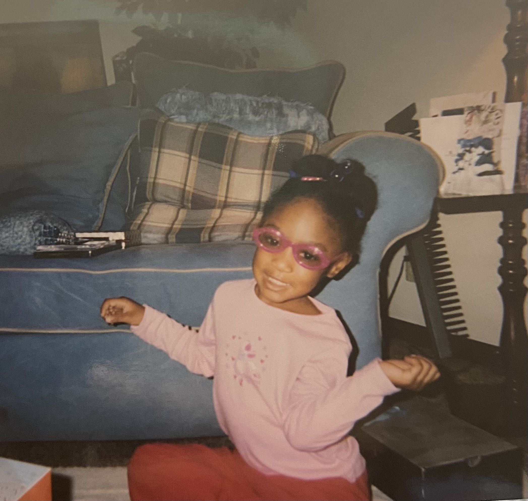 Child with pink glasses and a pink shirt sitting on the floor in a living room, smiling at the camera.