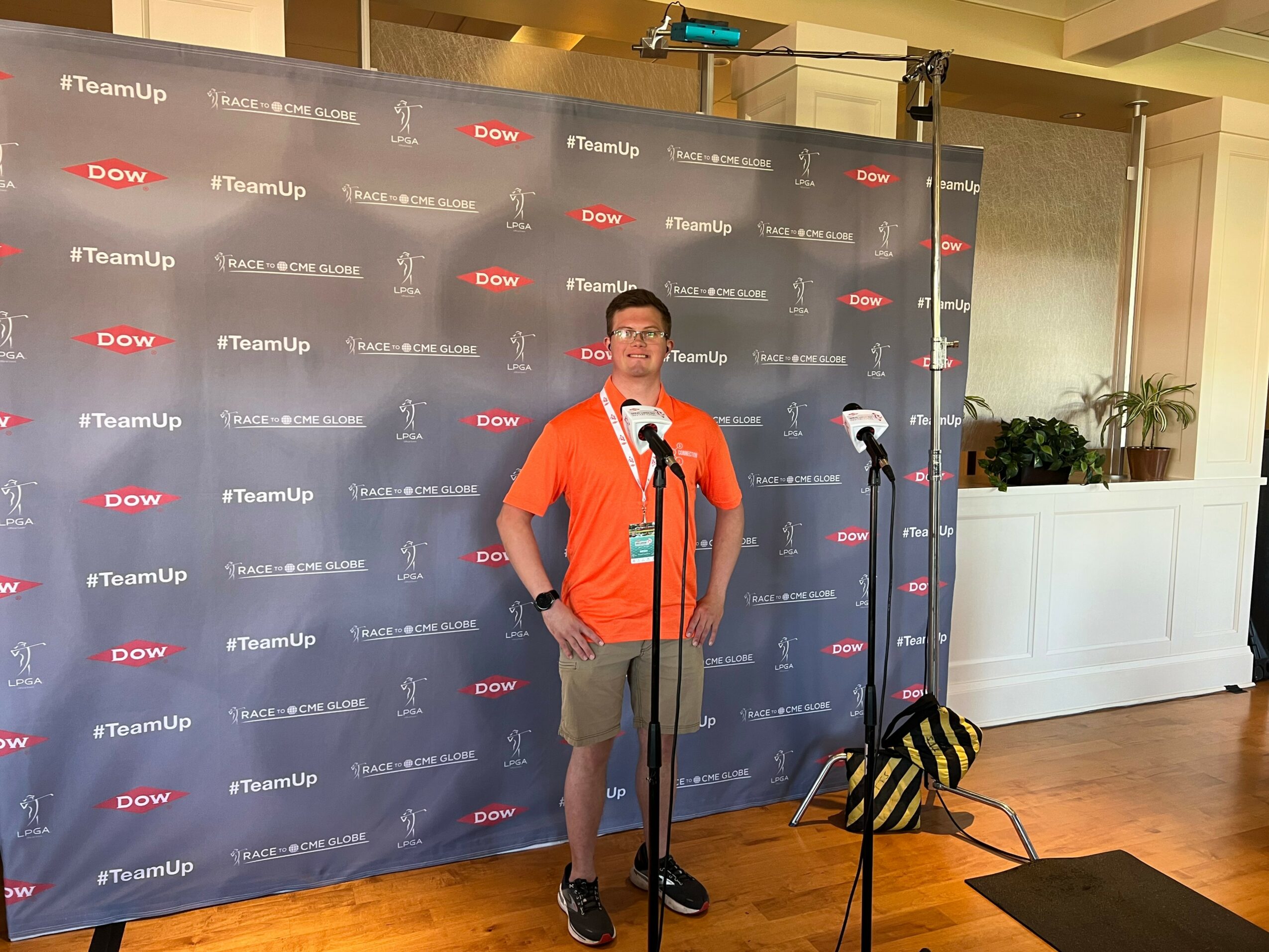Young man in orange shirt and shorts standing in front of a backdrop with logos, speaking into a microphone.