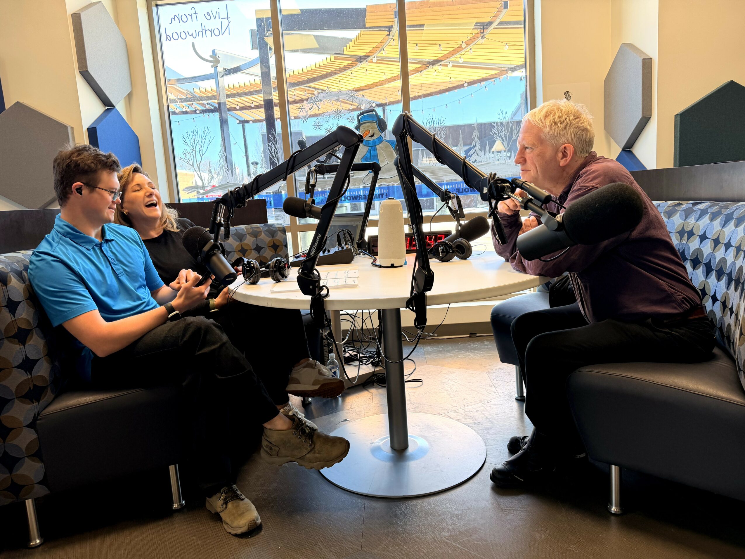 Four people sit around a table with microphones, engaging in a conversation in a bright room with large windows.
