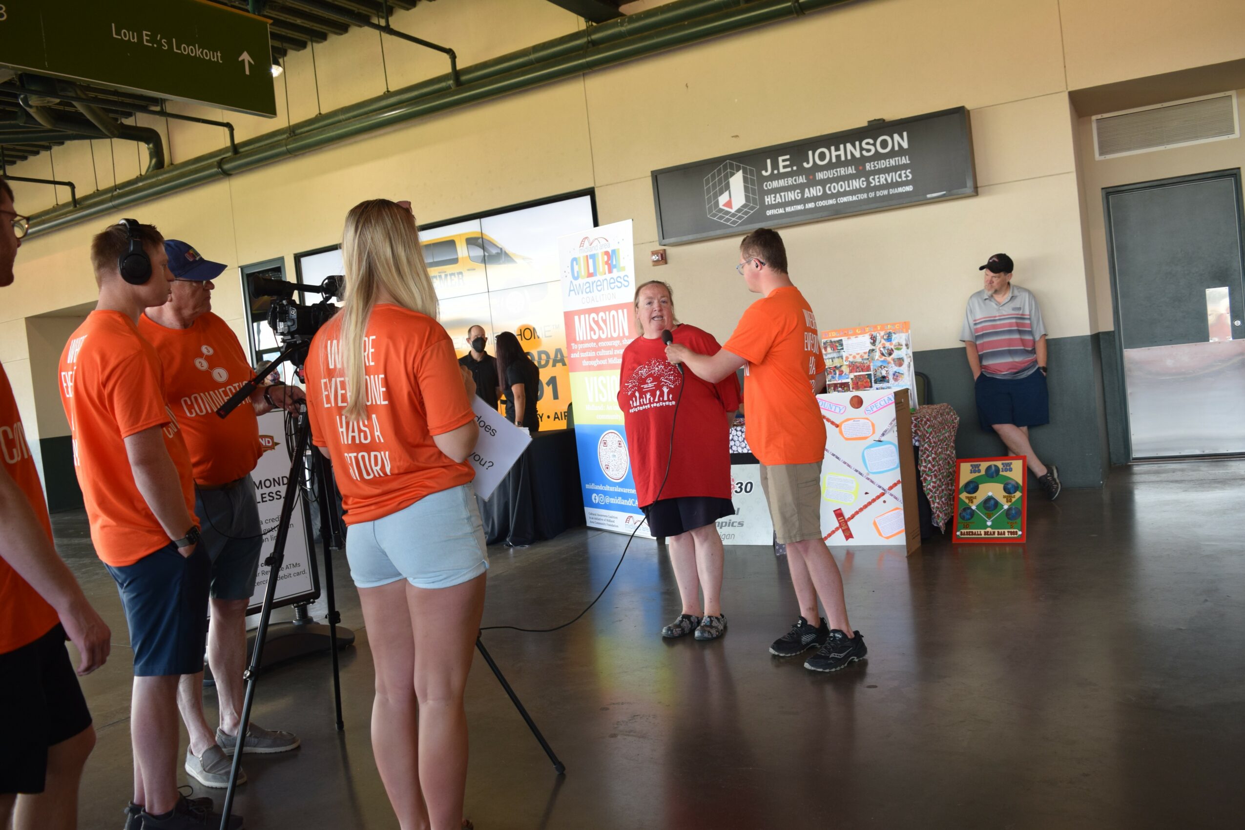 Group of people at an indoor event, some wearing orange shirts, with a woman receiving a red shirt from a man, and others standing nearby.