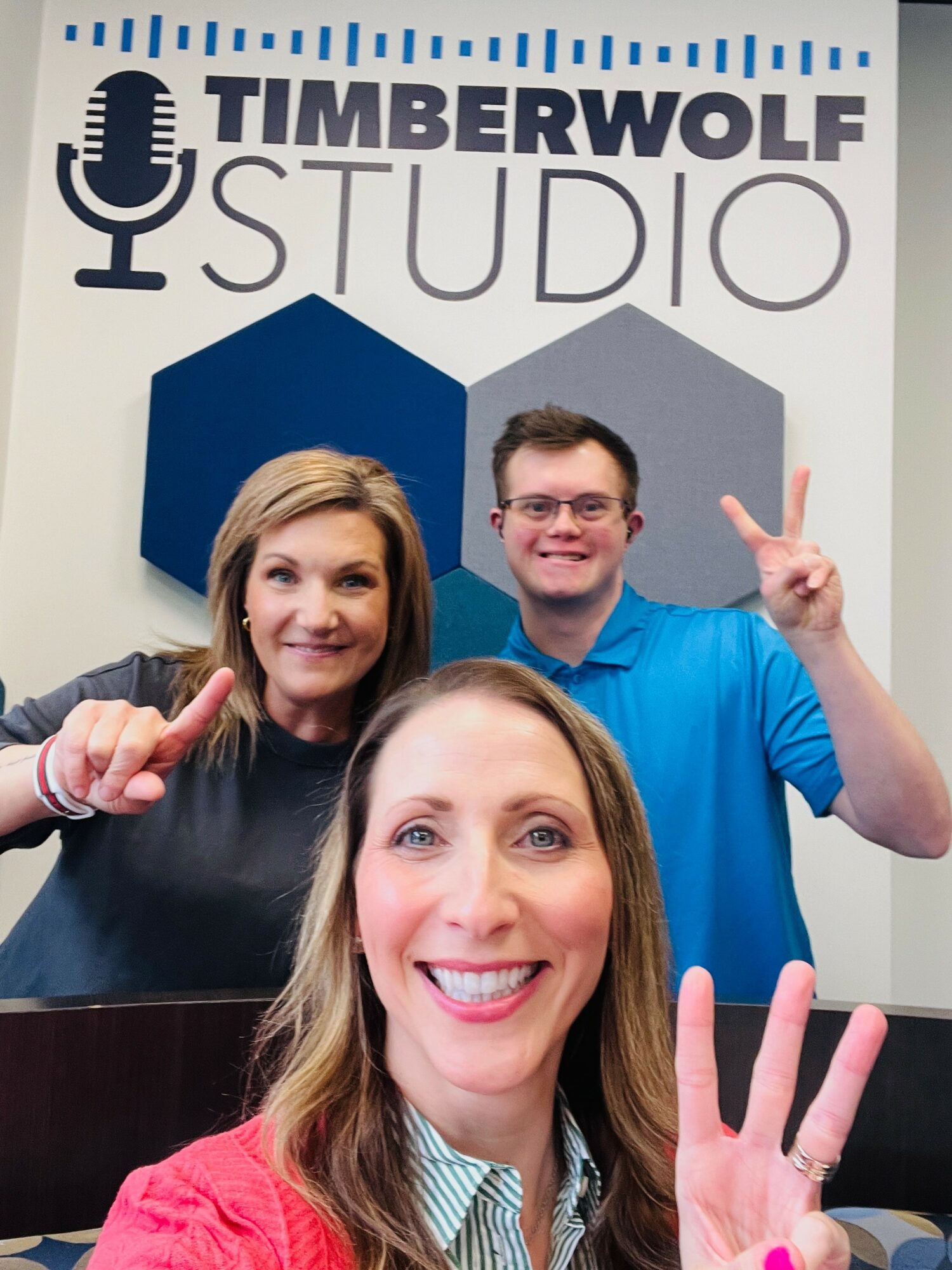 Three people smiling and making peace signs in front of a Timberwolf Studio backdrop with blue and gray hexagons.