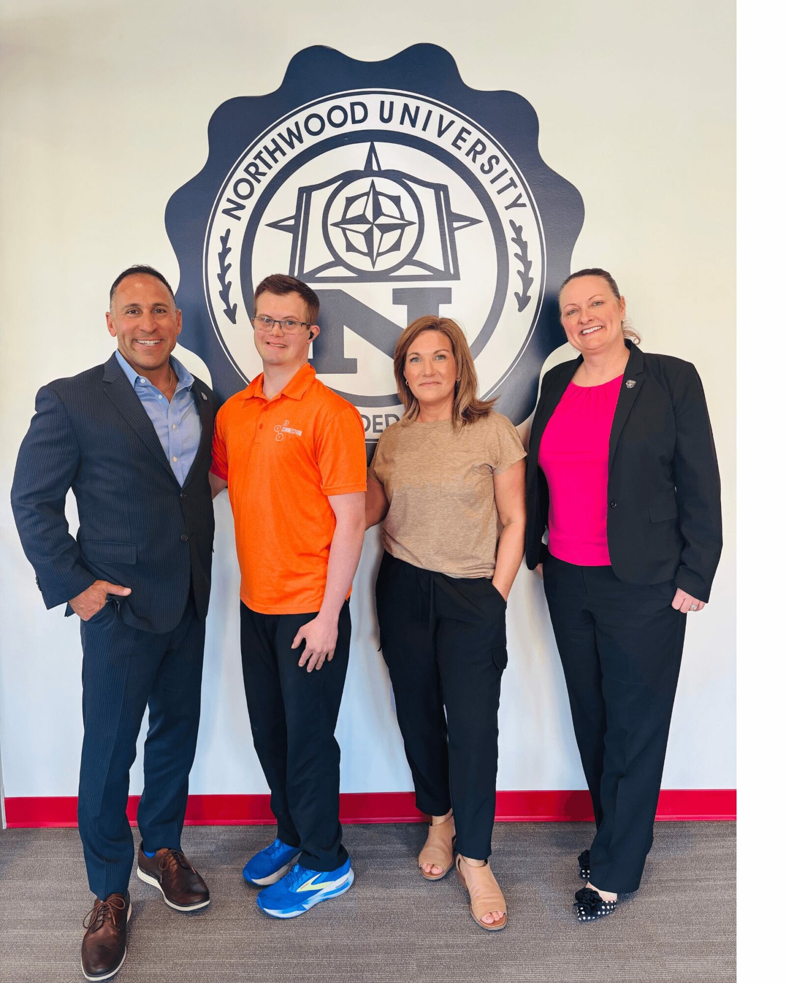 Four people standing in front of Northwood University logo, smiling, indoors, casual and business attire, group photo.