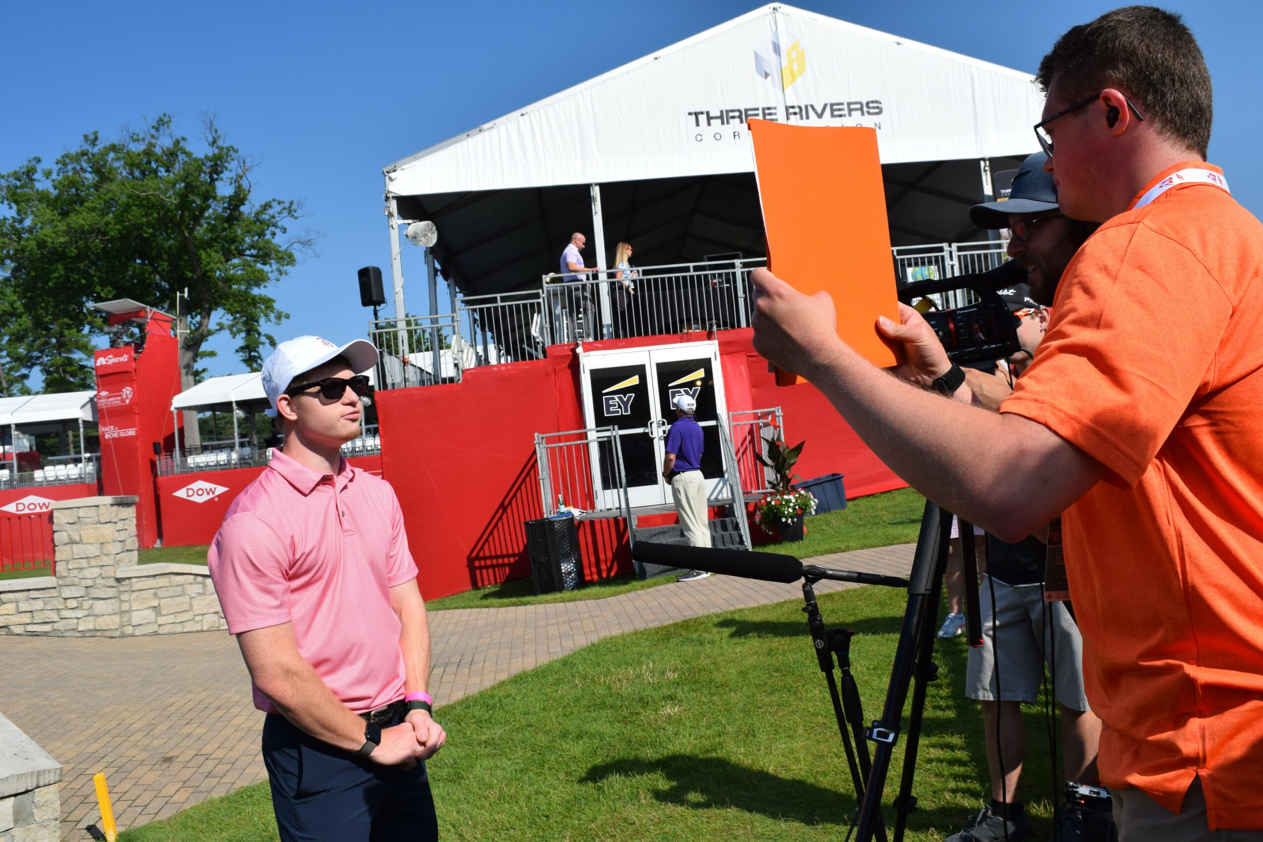Two men are filming at an outdoor event with a stage and red structures in the background, under a clear blue sky.