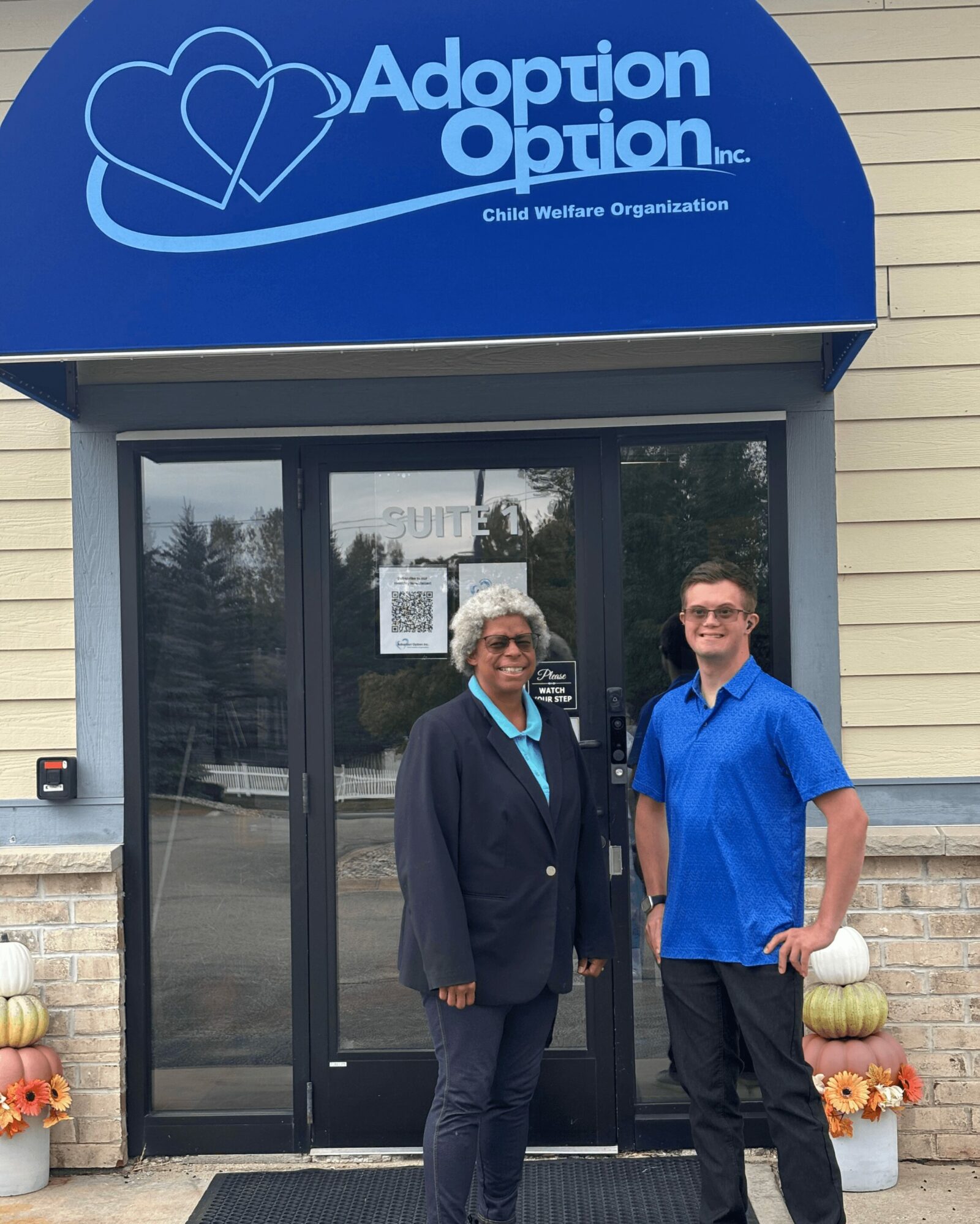 Two people standing outside Adoption Option Inc. building, smiling, with pumpkins and flowers nearby.