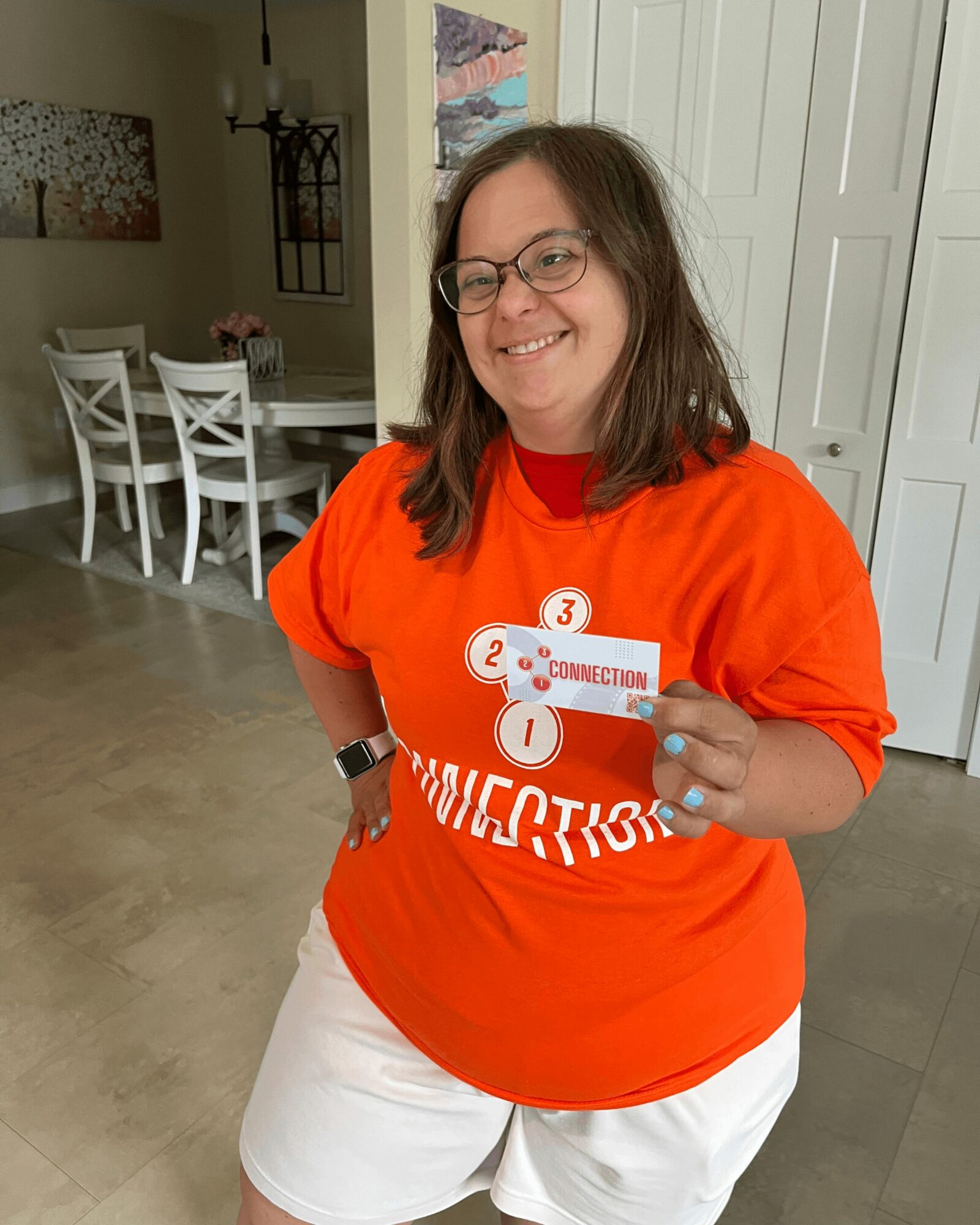 Woman with glasses smiling, wearing an orange shirt and white shorts, holding a card, standing indoors.