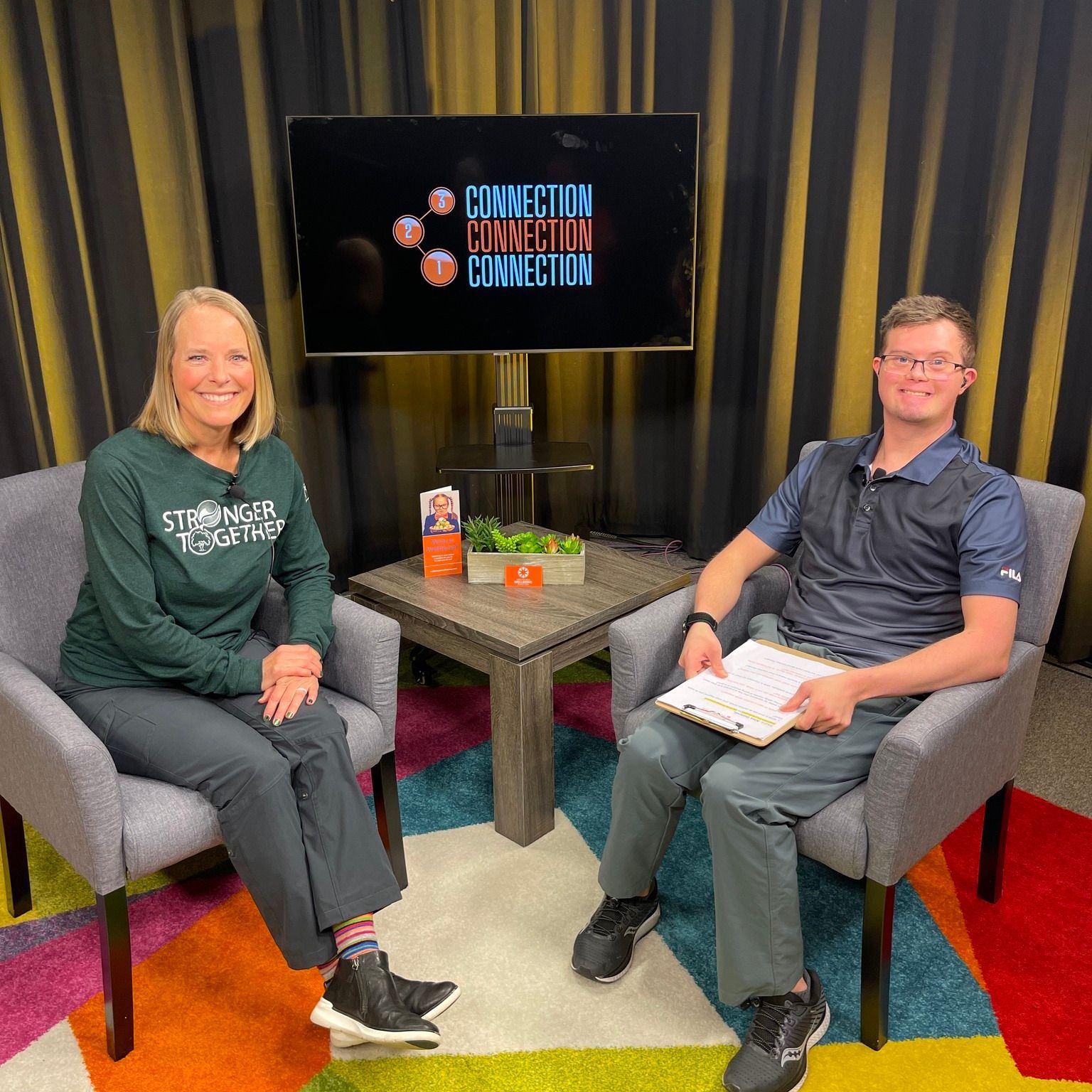 Two people sitting in chairs with a table between them, a screen behind displaying the word 'CONNECTION' three times, colorful carpet, and curtains.