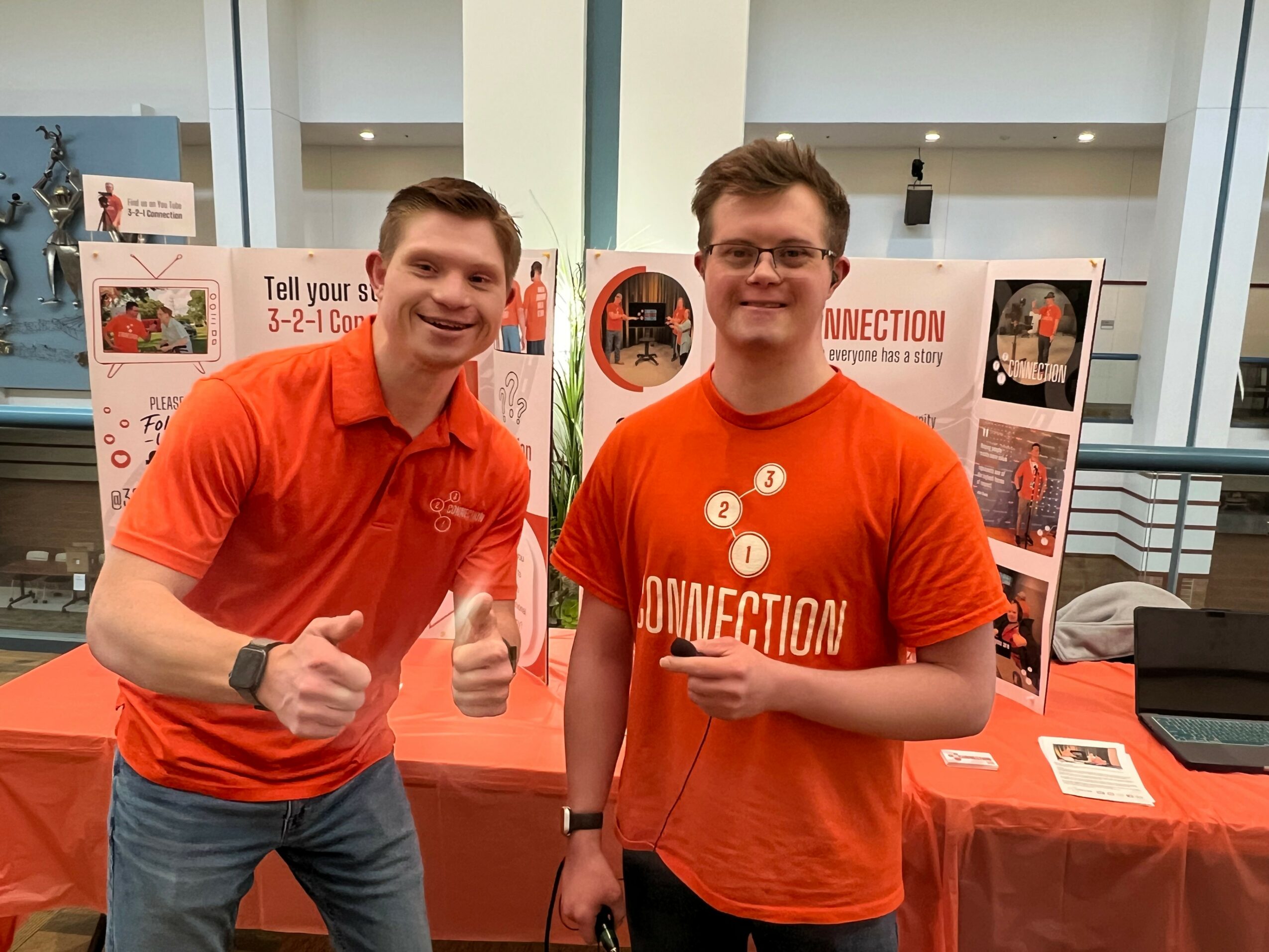 Two young men in orange shirts standing in front of display boards at an indoor event, smiling and giving thumbs up.