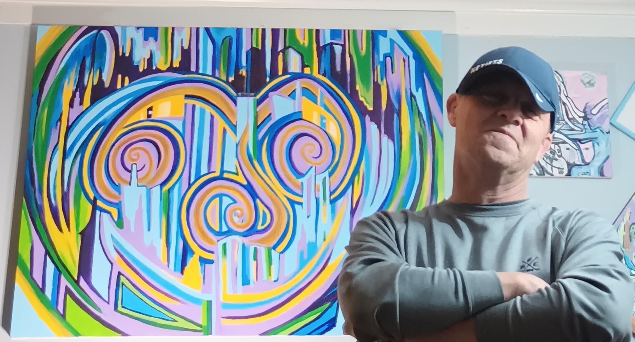 Person wearing a cap and gray shirt standing with arms crossed in front of colorful abstract artwork.
