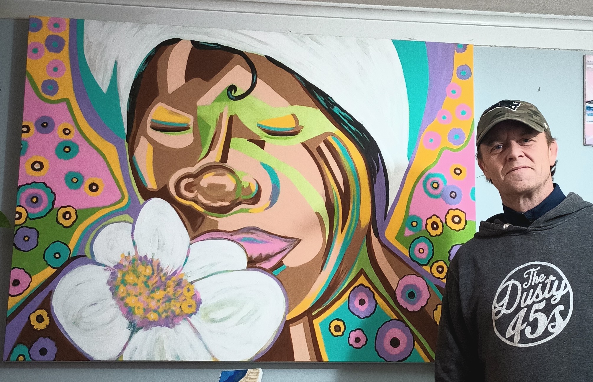 Man standing next to colorful mural of a woman's face with a flower, vibrant patterns, and bright colors.