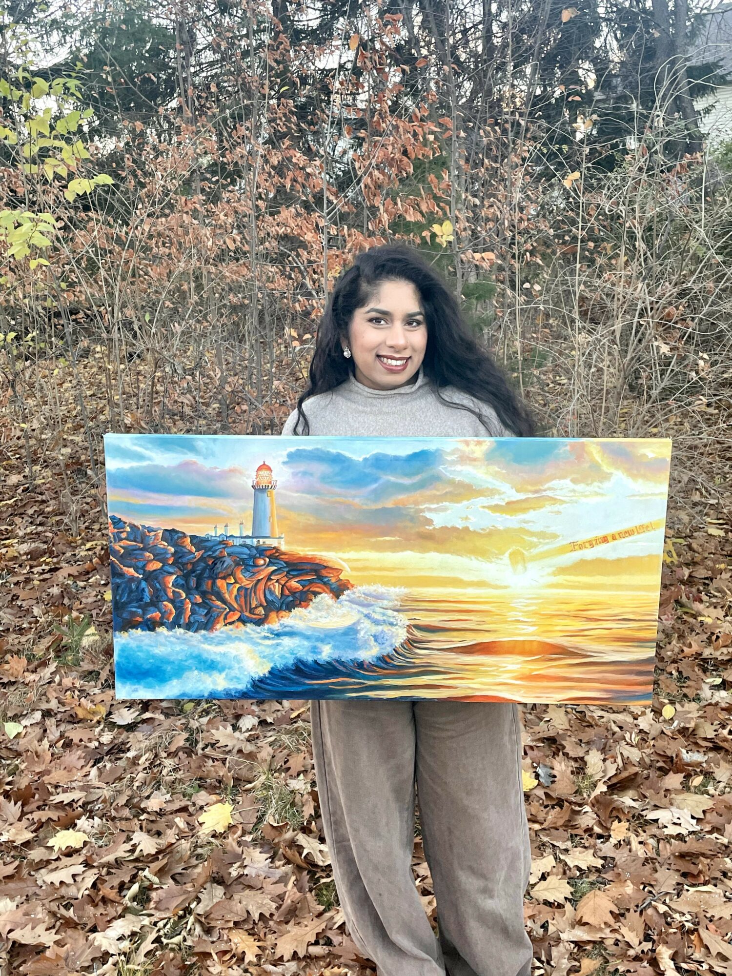 Woman holding a large painting of a lighthouse and sunset over the ocean outdoors, surrounded by trees and fallen leaves.
