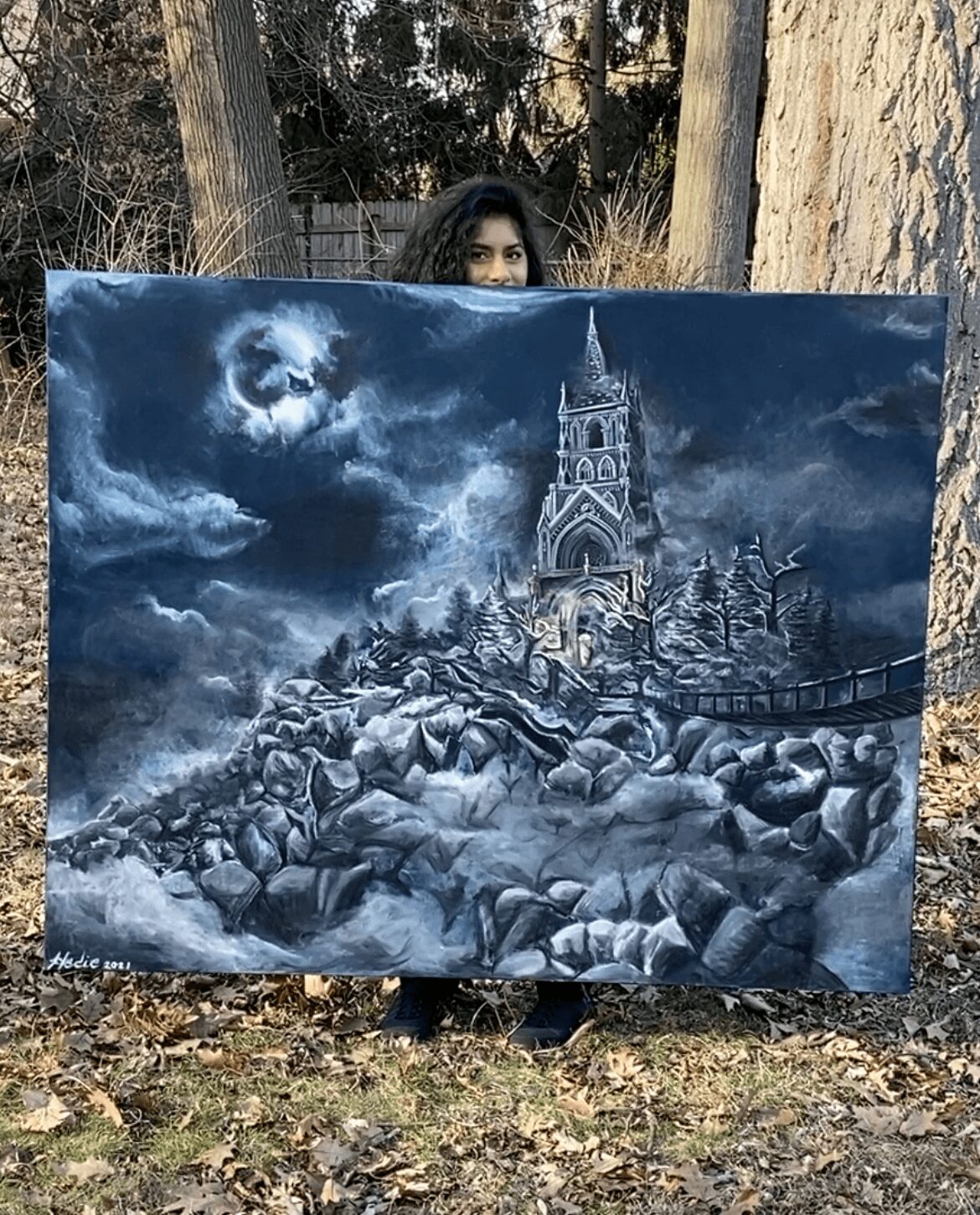 Person holding a painting of a moonlit, snowy landscape with a church and bridge, trees, and rocks, outdoors in a wooded area.