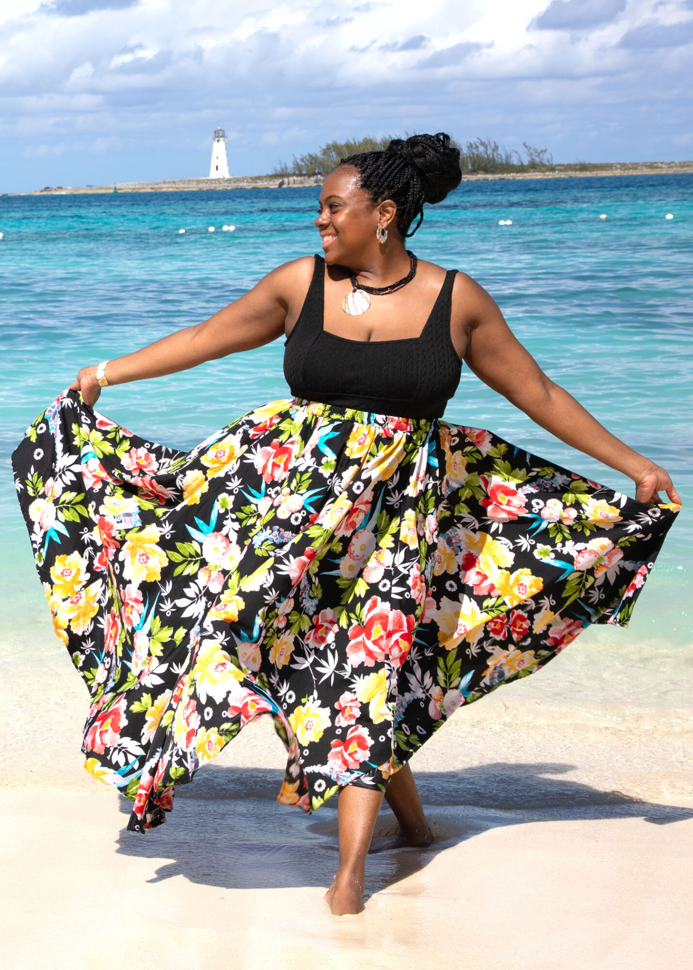 Woman in a black top and floral skirt standing on beach, holding skirt out, ocean and lighthouse in background.